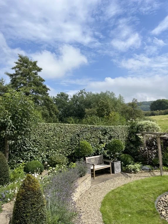A well-maintained garden with a gravel pathway, green grass, a wooden bench, a small cabinet, and various bushes and plants under a partly cloudy sky.