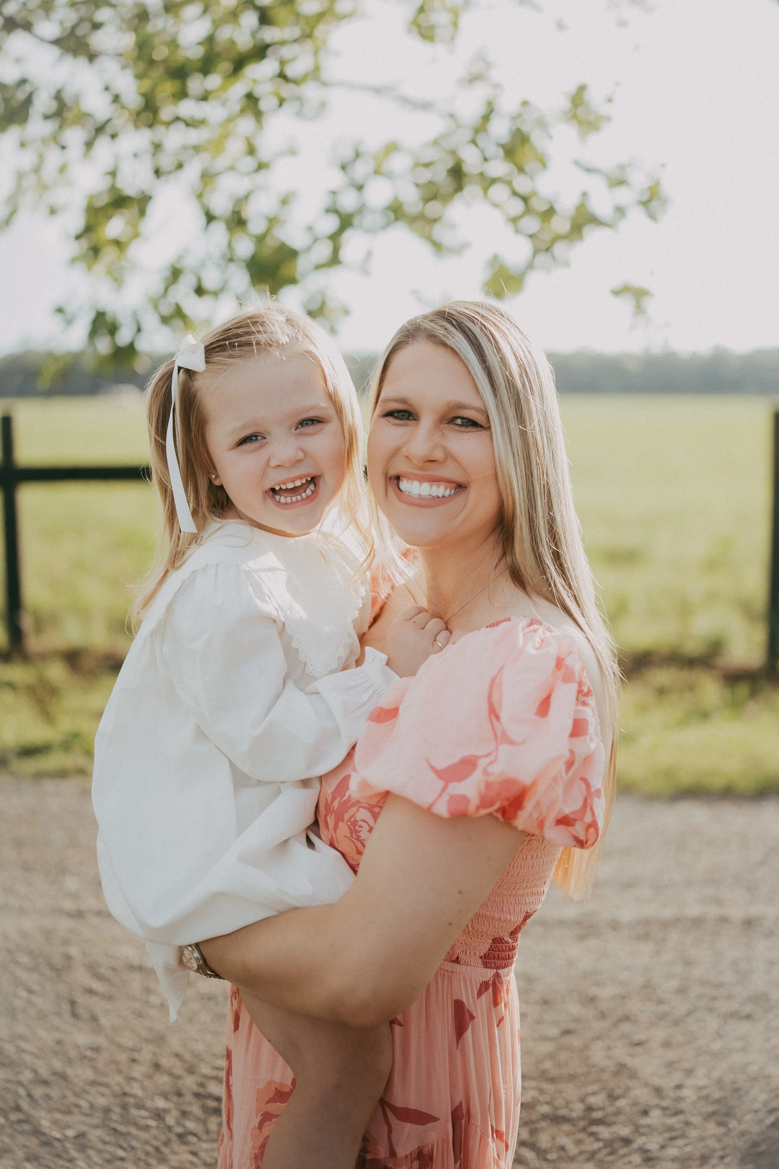 A mother holds her smiling daughter close as they share a joyful embrace beneath sunlit oak trees, creating a timeless outdoor family portrait filled with laughter, love, and genuine connection.