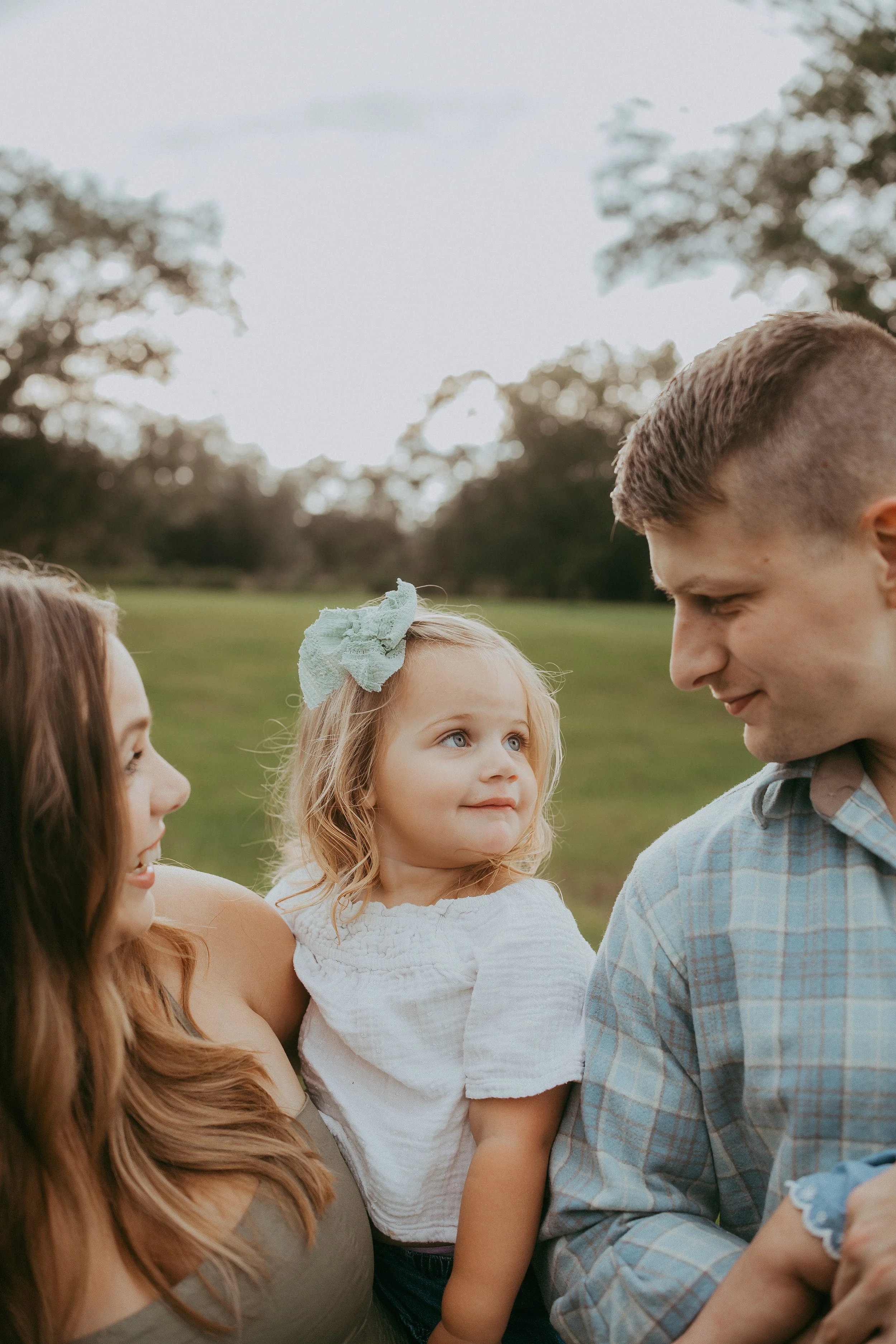 A little girl with a soft blue bow looks lovingly between her mother and father as they smile back at her, capturing a quiet family moment filled with warmth, trust, and connection in an open grassy field.