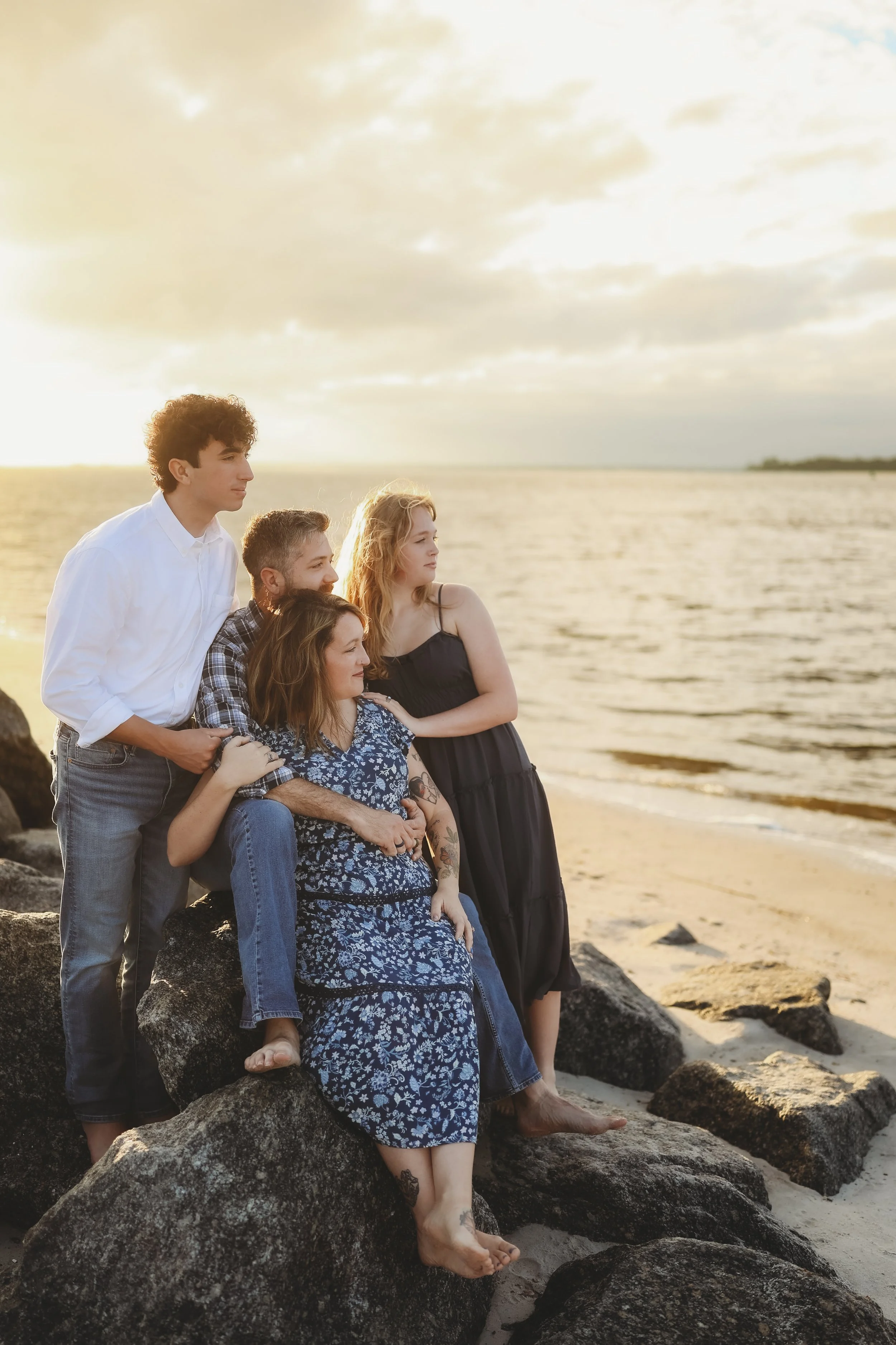 A family sits together on the shoreline rocks, watching the sunset over the water during a peaceful beach portrait session.