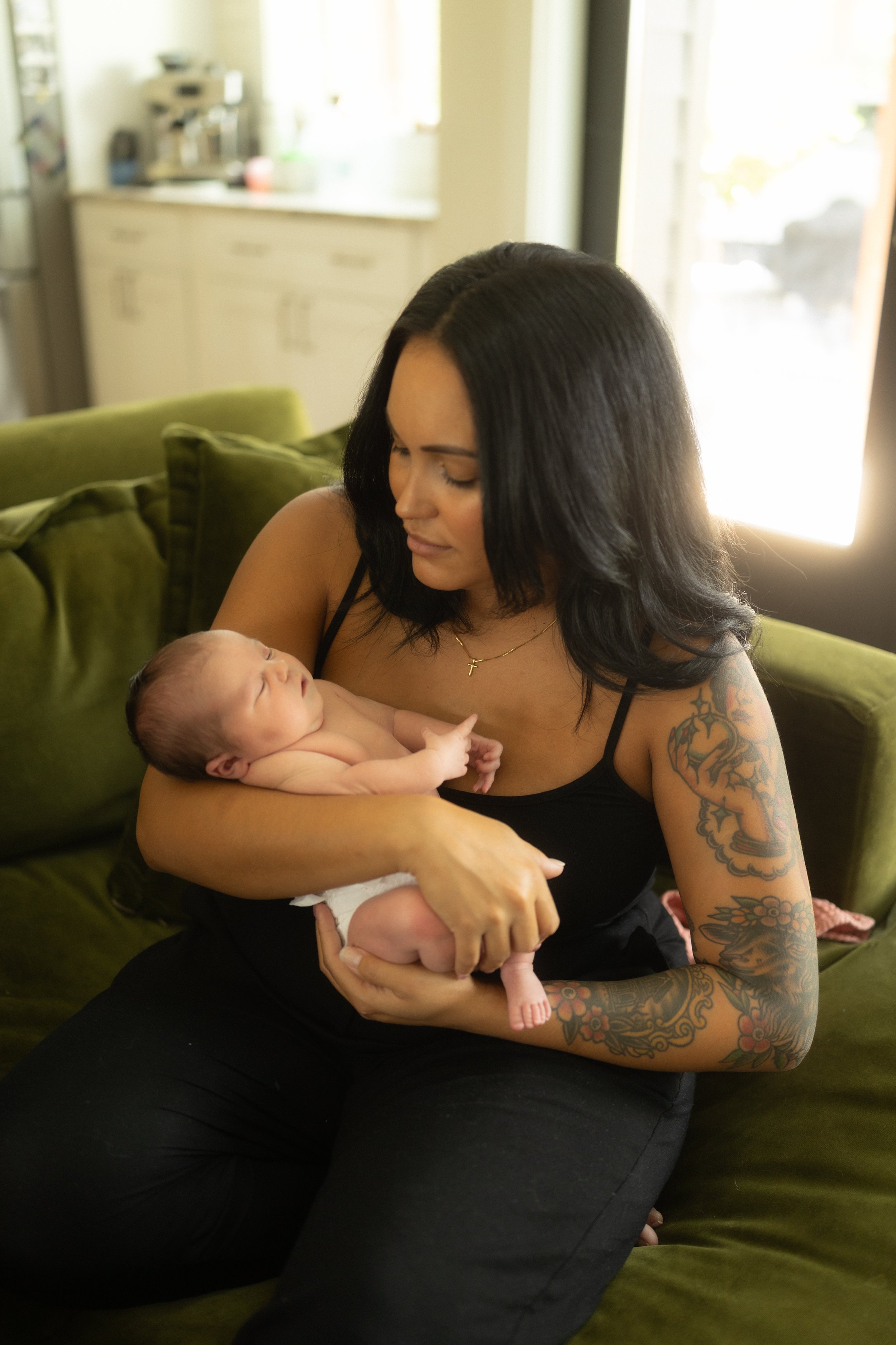 Mother cradles her newborn on a cozy living room sofa, gazing down with a gentle smile during a relaxed in-home lifestyle newborn photography session that captures the beauty of everyday connection.
