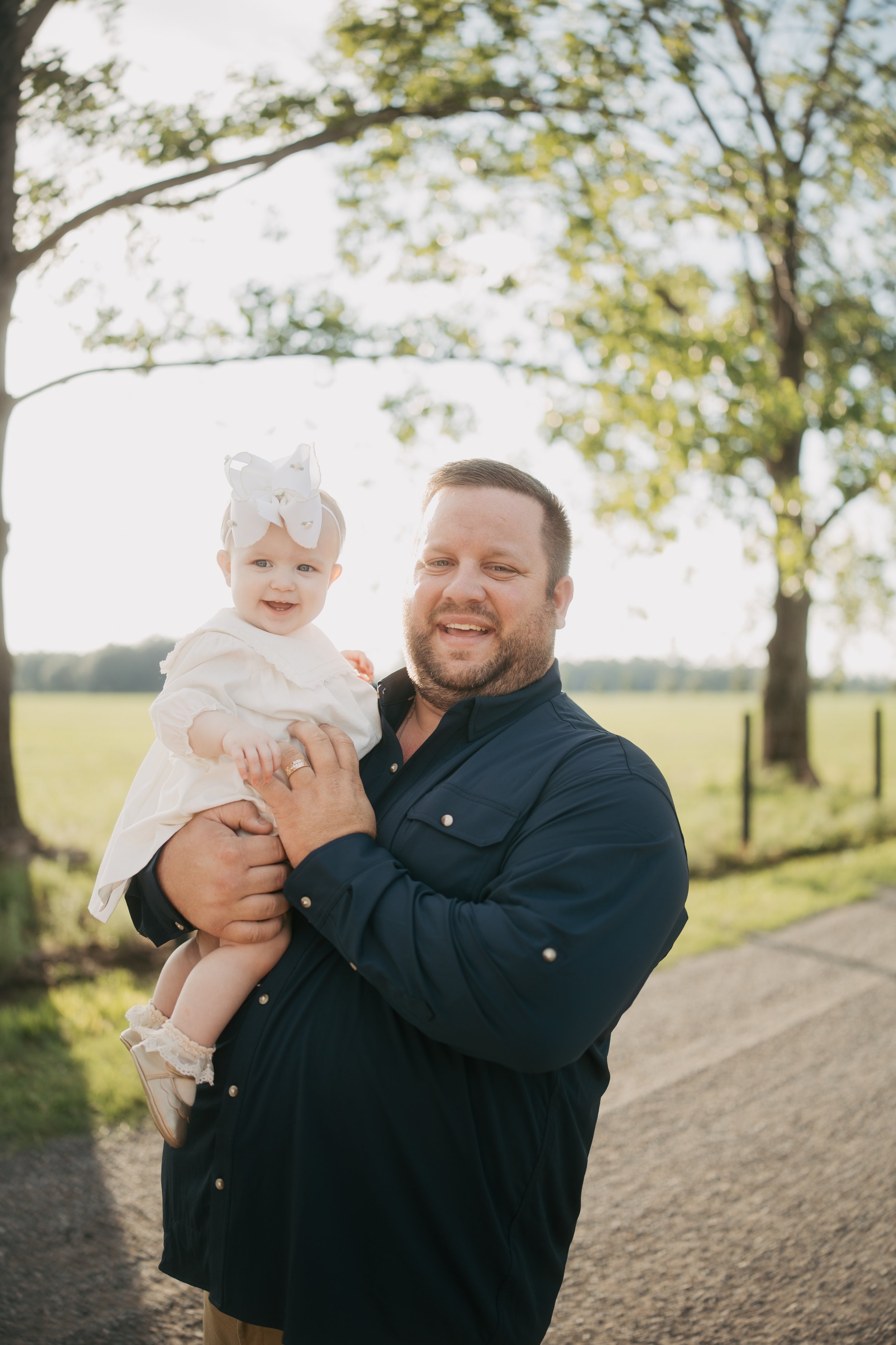A father smiles as he holds his baby daughter in his arms beneath towering oak trees, capturing a tender father-daughter moment during a warm, sunlit family session in the Florida countryside.