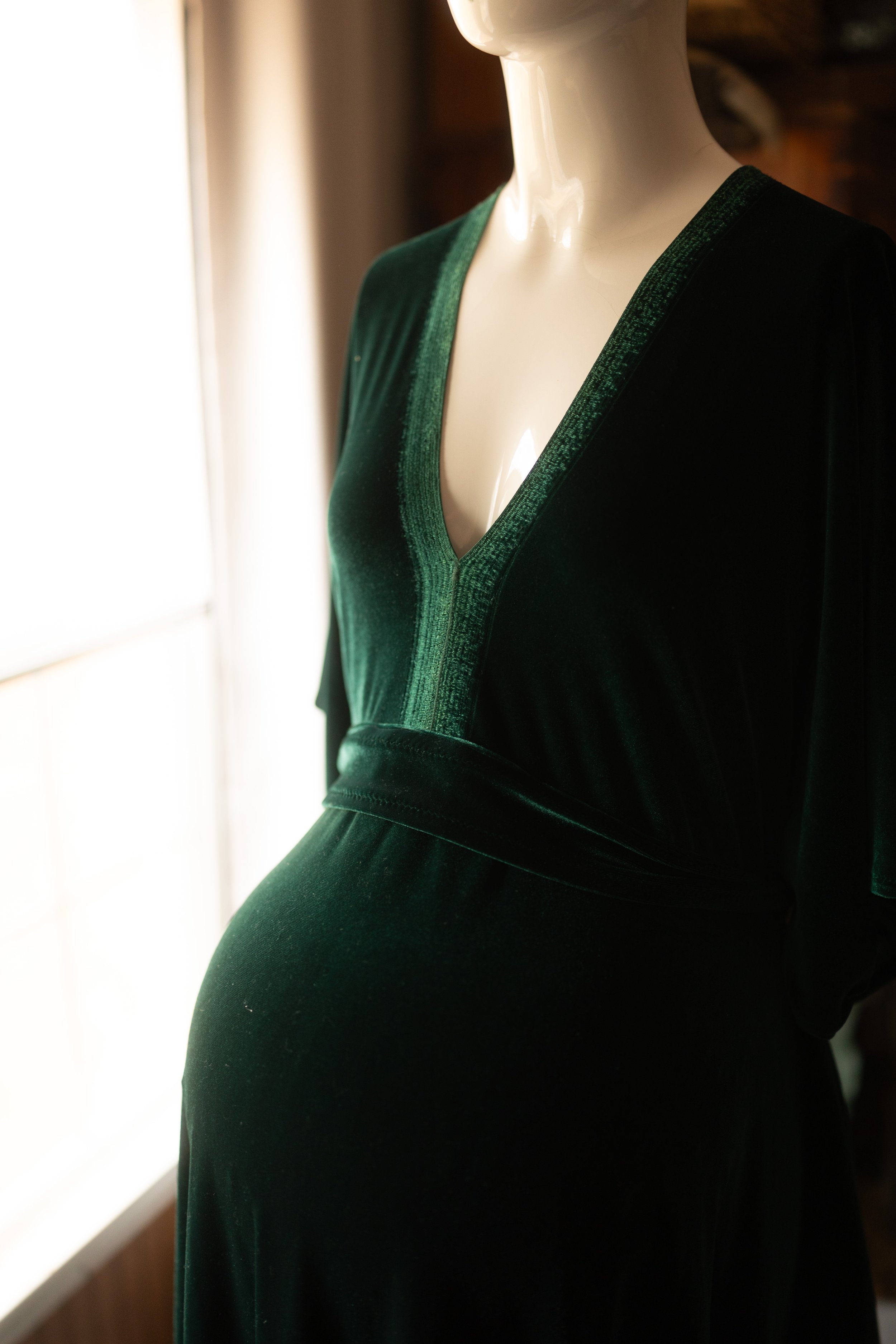 Deep emerald velvet maxi dress with a wrap-style V-neck bodice, long sleeves, and a flowing skirt, styled on a mannequin in soft natural light.