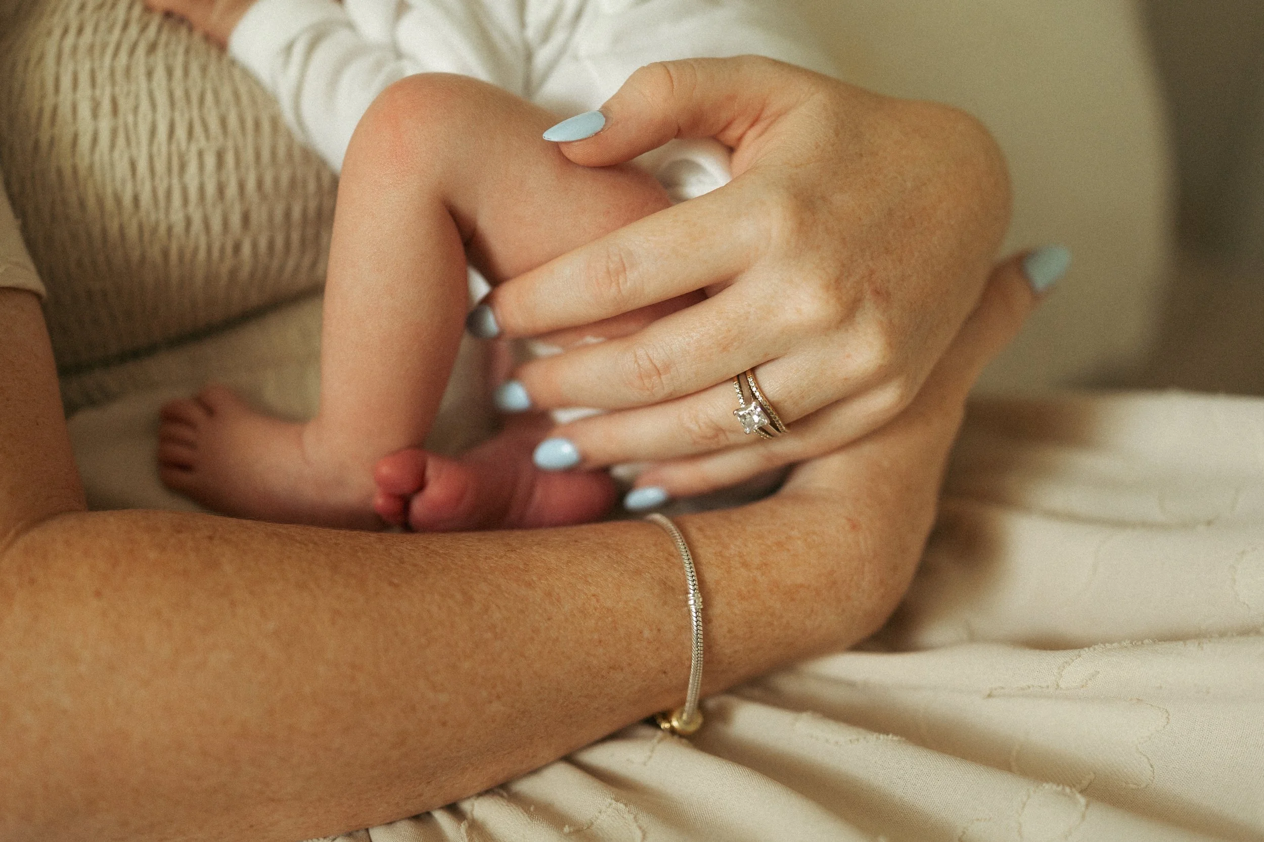Close-up of a mother's hands gently holding her newborn, highlighting tiny baby feet, delicate fingers, and a quiet moment of love during an in-home lifestyle newborn photography session.