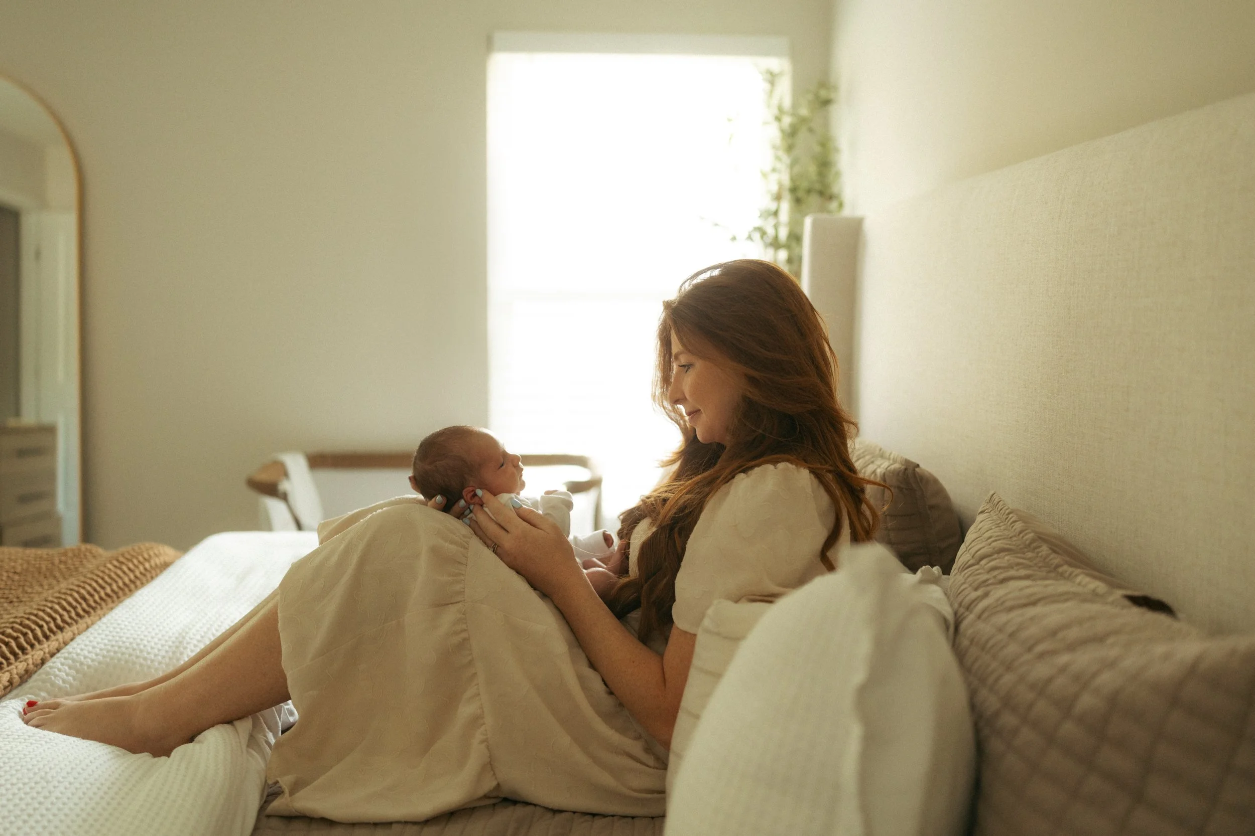Mother smiles softly at her newborn while sitting on a bed in a sunlit bedroom, sharing a tender face-to-face moment during an intimate in-home lifestyle newborn photography session.