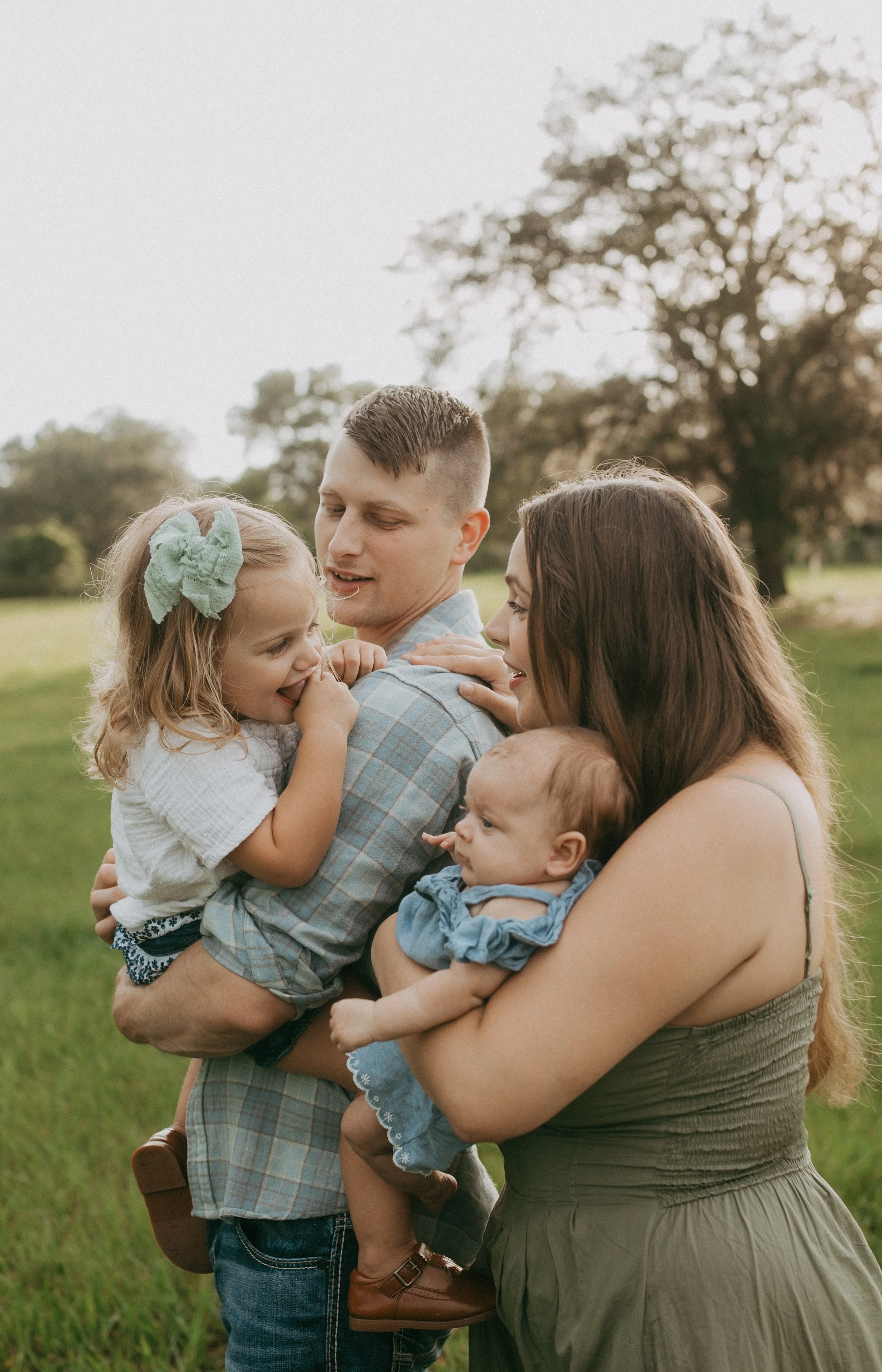 A mother and father cuddle their baby and toddler together in a grassy field as their young daughter smiles shyly in her father's arms, capturing a tender family moment filled with love and connection.