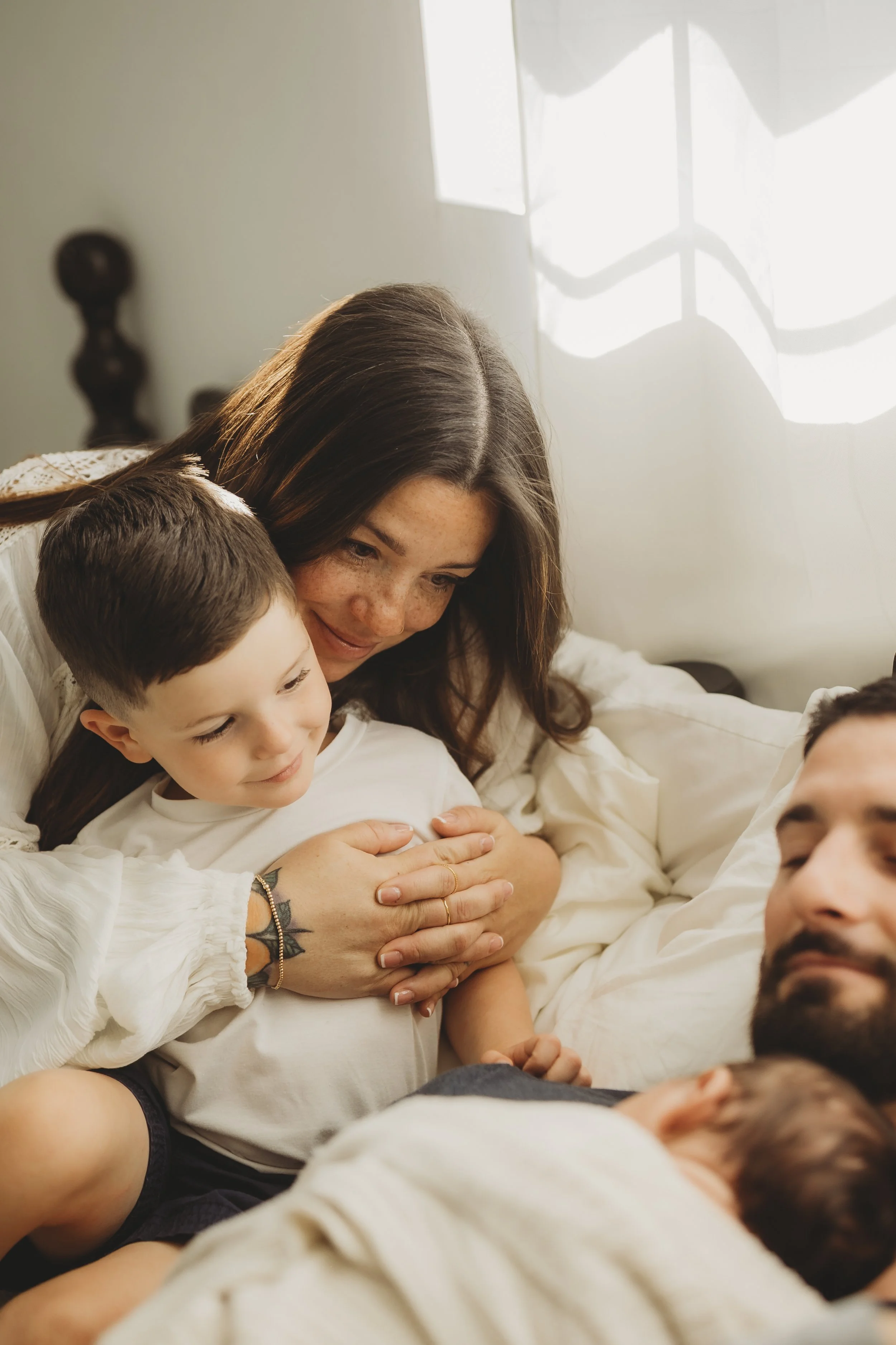 Mother resting on a bed with her child held close while watching the newborn sleep nearby in soft window light.
