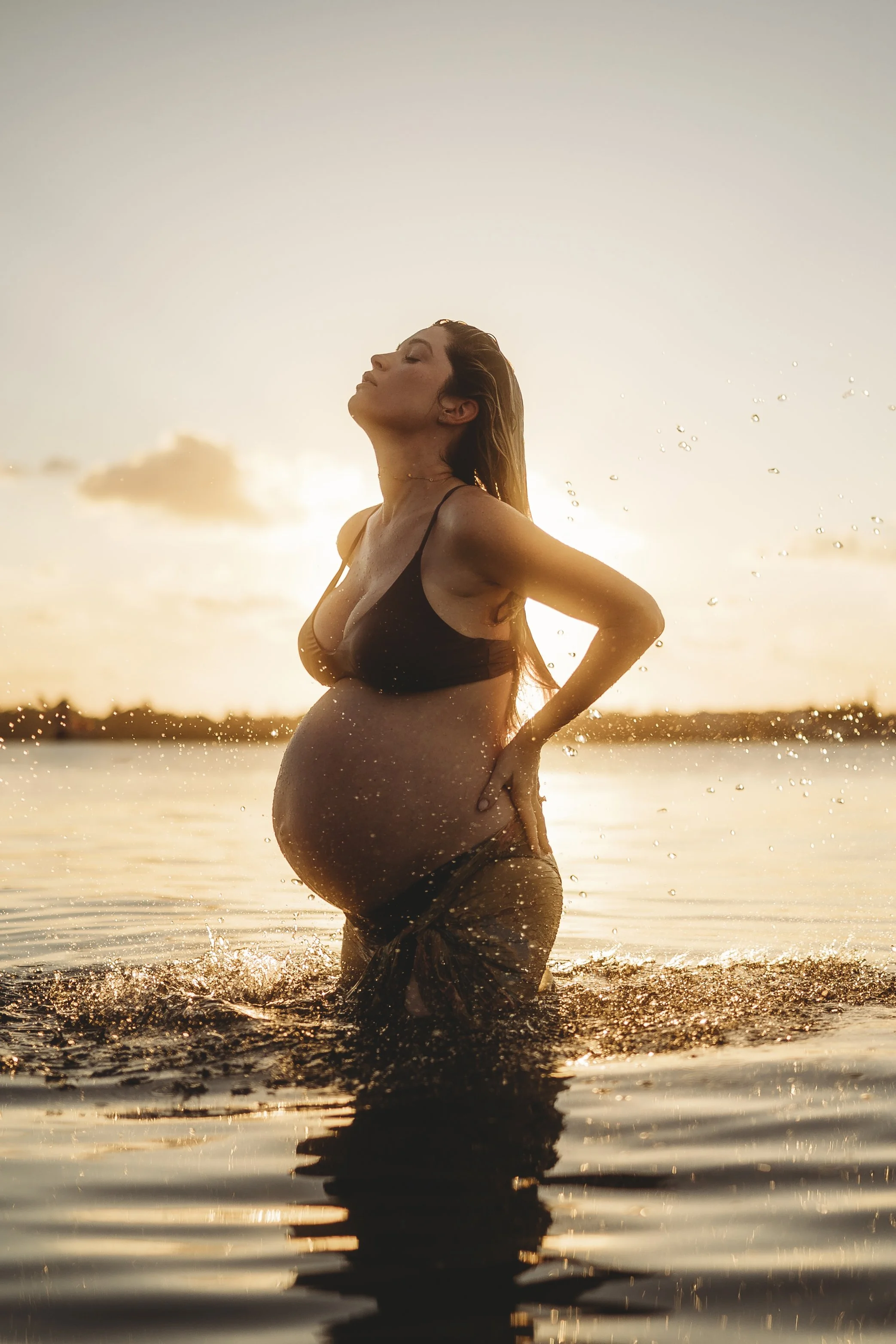 Fine art maternity image of an expectant mother in water at golden hour, her silhouette glowing as sunlight reflects across rippling waves.