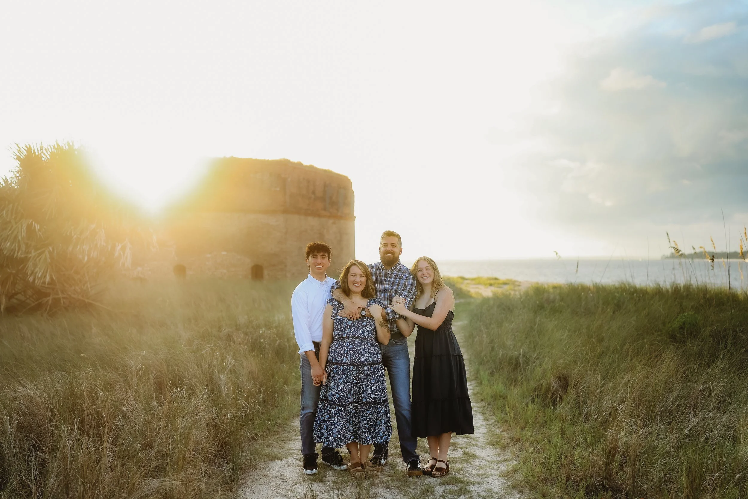 A family of four smiles together on a sandy path with the historic fort glowing behind them at sunset.