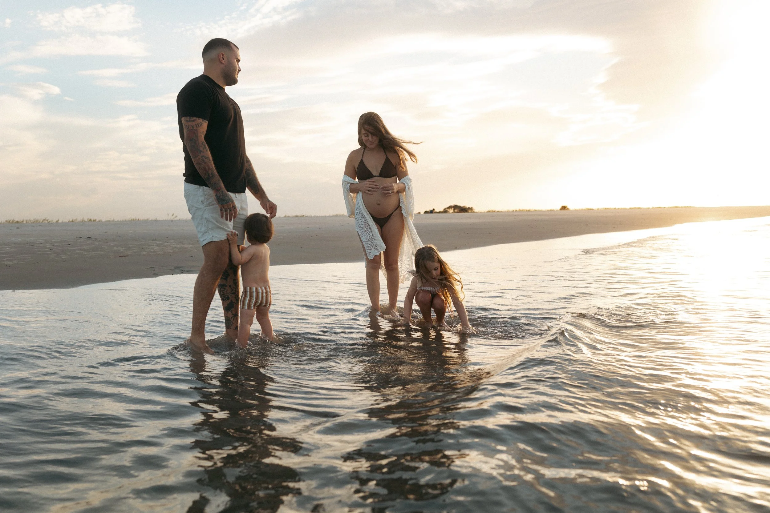 Pregnant mother stands in shallow water with her family at sunset on Amelia Island Beach as two young children play in the gentle waves.