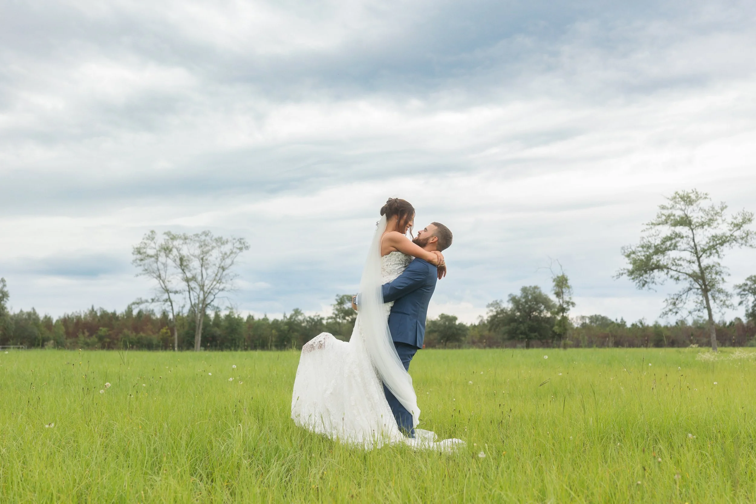 Emma wraps her arms around Dylan as he lifts her in a green field beneath a dramatic cloudy sky at Red River Estate in Waynesville, Georgia.