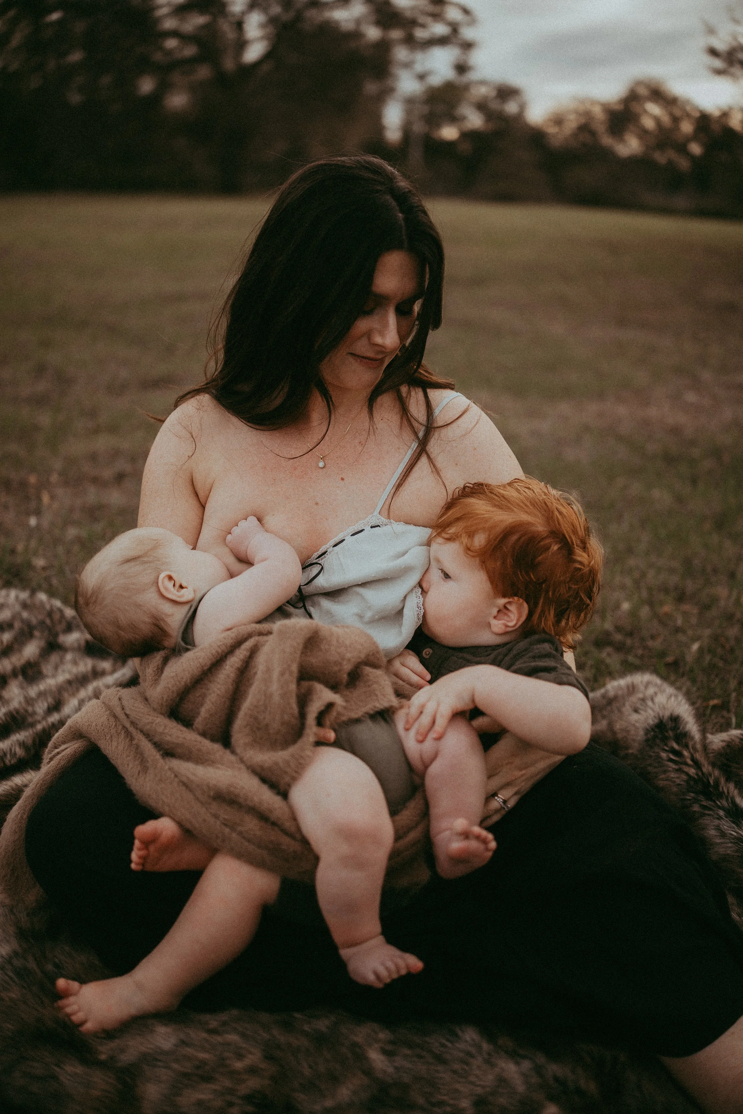 A woman with dark hair wearing a white tank top and necklace, sitting outdoors on a blanket with two young children, one with red hair and one with blonde hair, holding and breastfeeding them, surrounded by natural scenery with trees and grass at dusk.