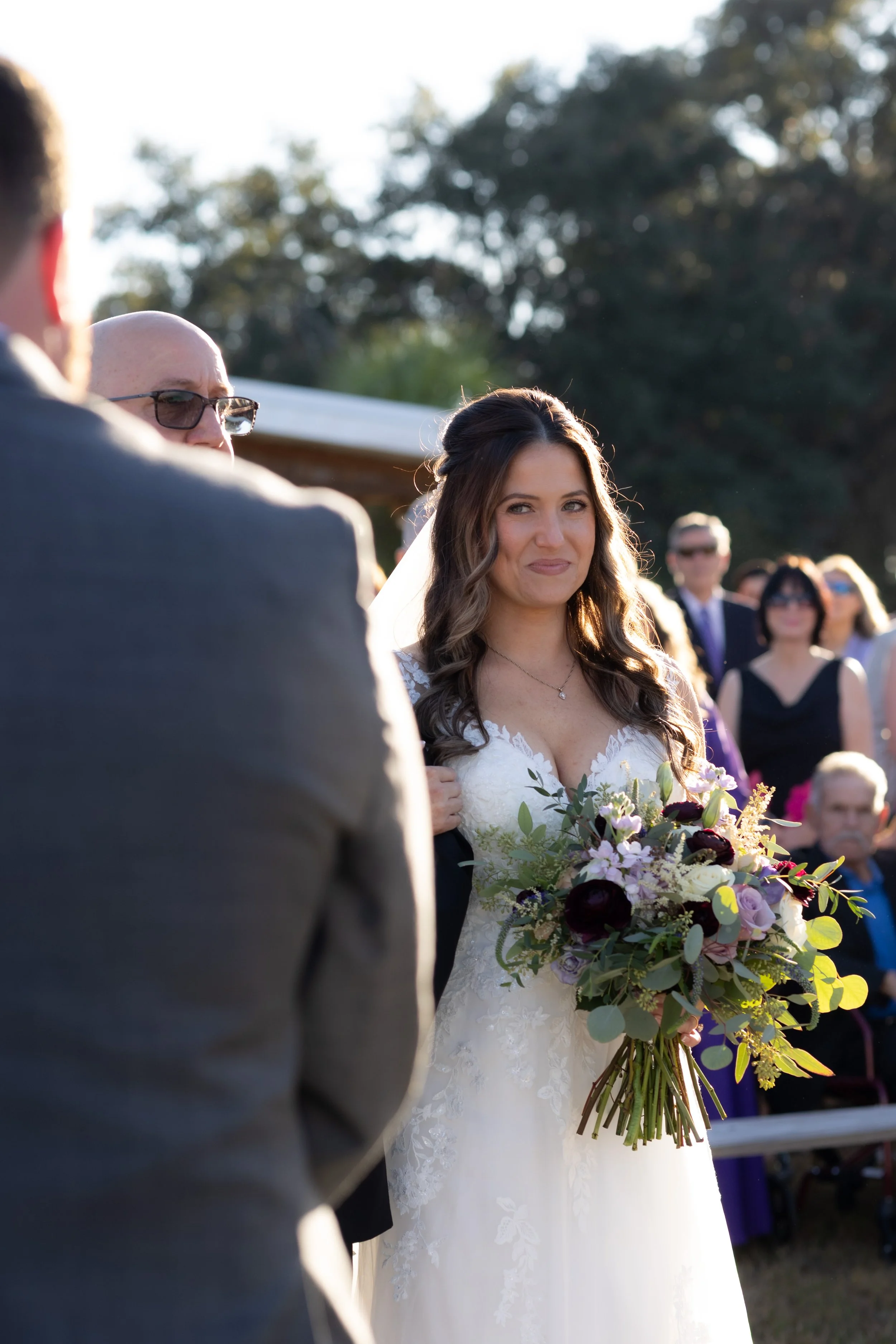 Bride smiling with emotion during her outdoor wedding ceremony, holding a lush bouquet as she faces her partner.