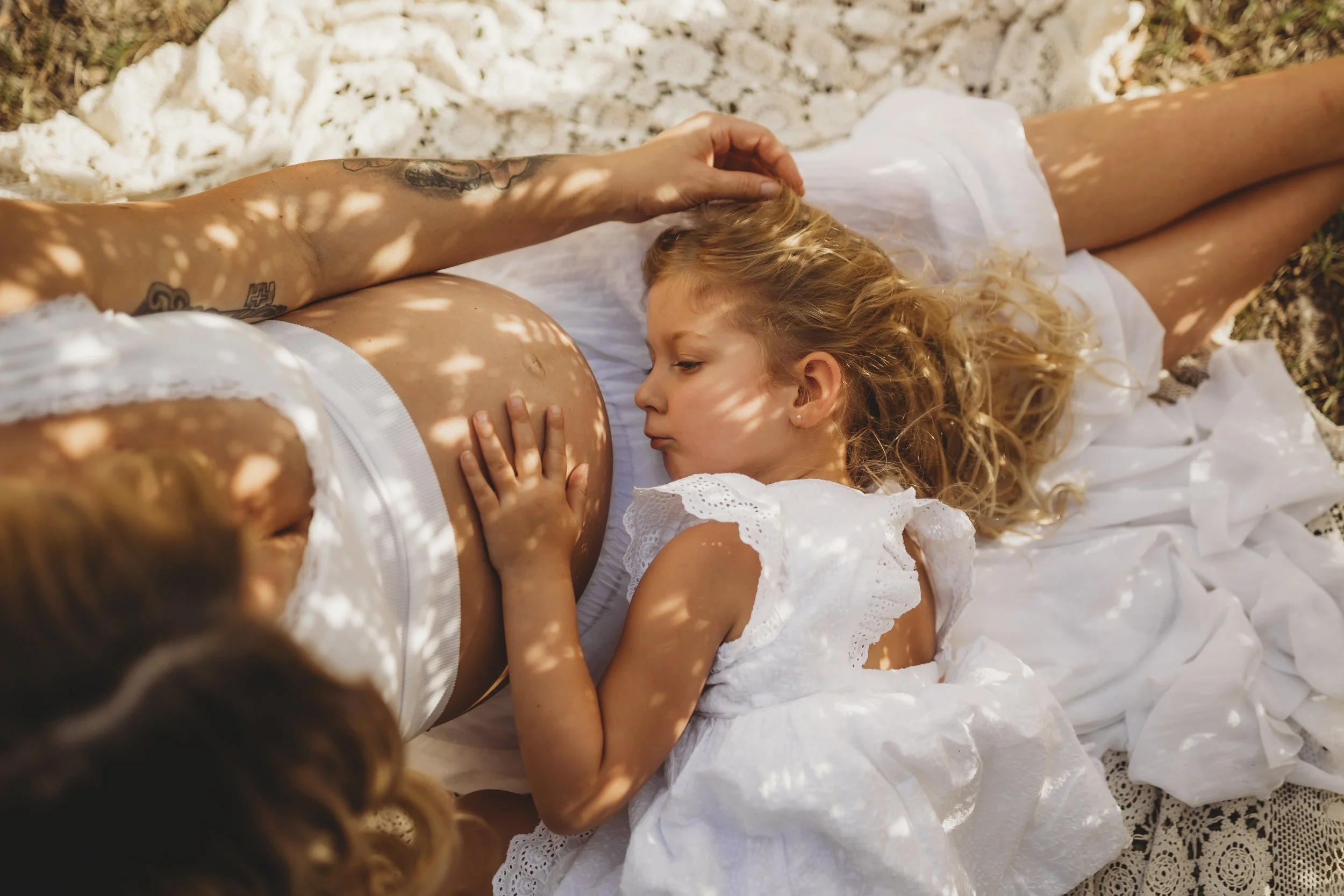 Dappled sunlight falls over a mother and child as little hands rest gently on a pregnant belly, capturing the beginning of sibling love.