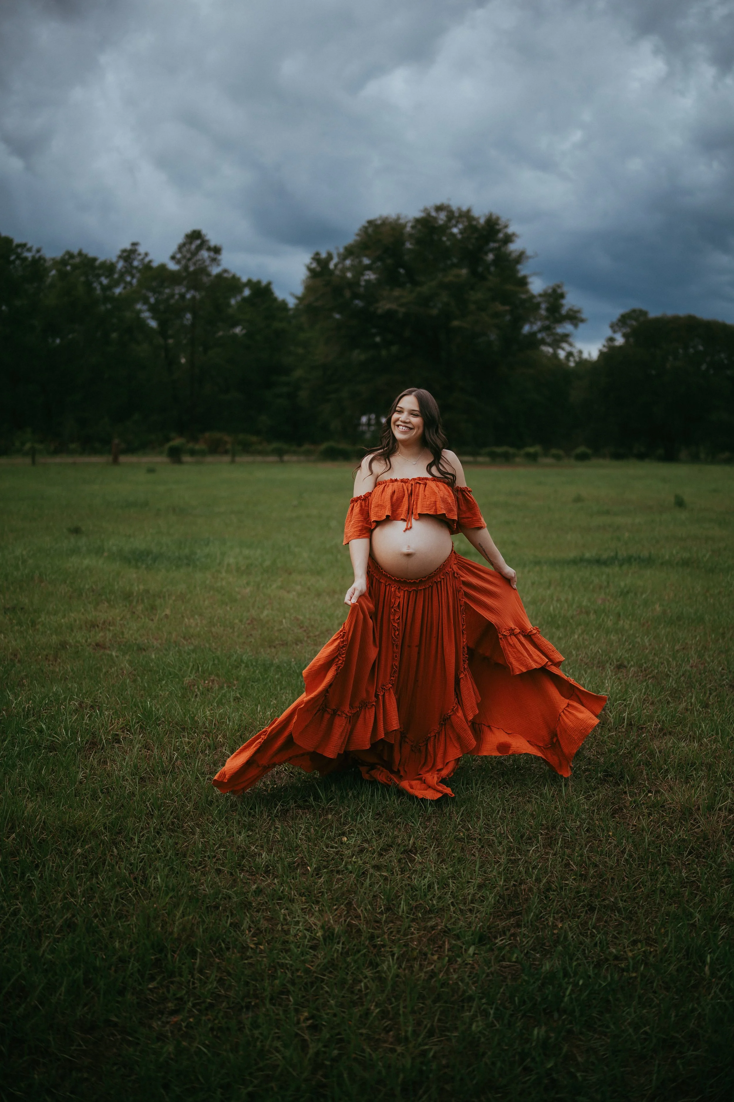 Expectant mother in a flowing rust-colored dress standing in a grassy field beneath dramatic storm clouds during a Hilliard, Florida maternity session.