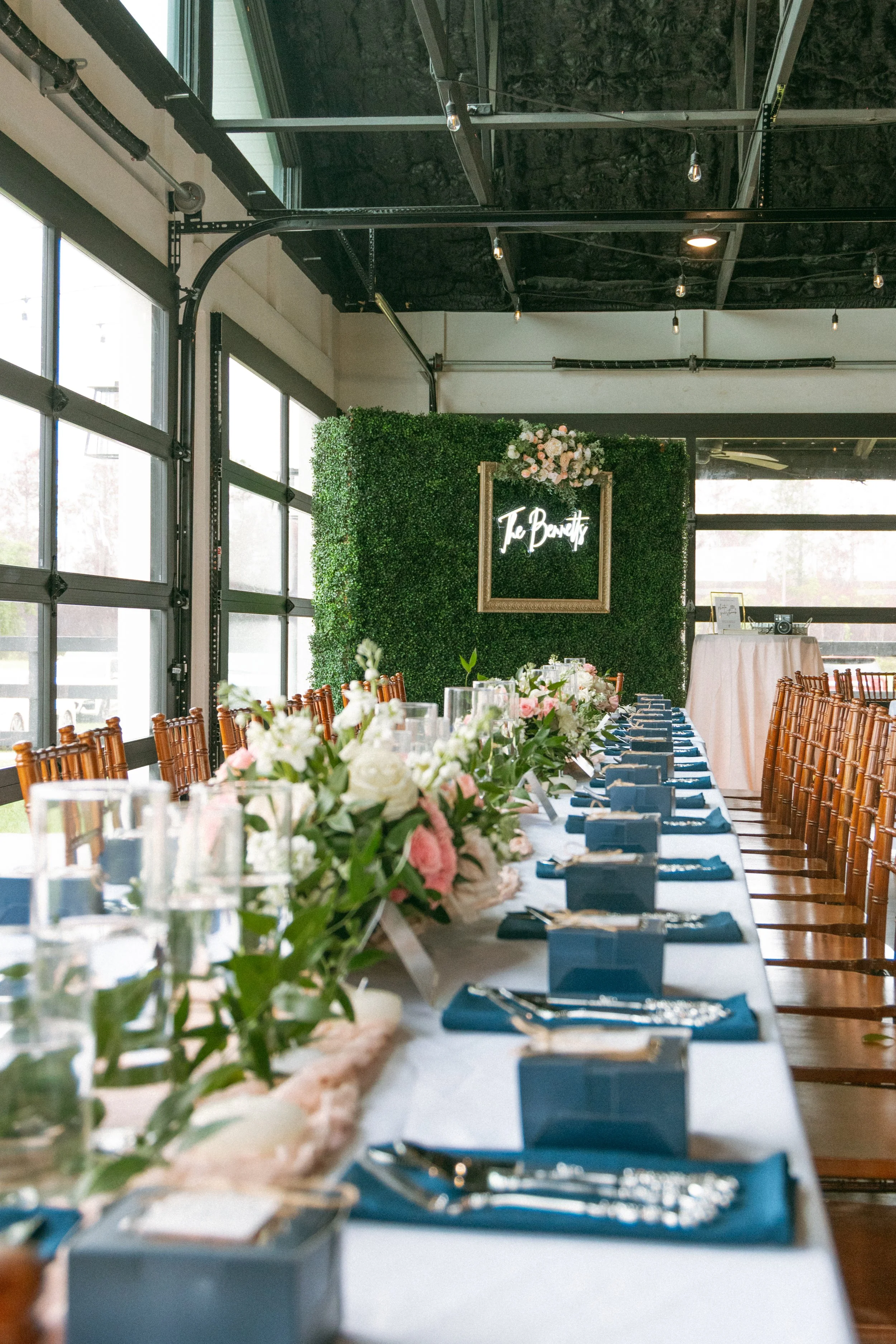 Wedding reception tables at Red River Estate decorated with blush and white flowers, navy napkins, candles, and a greenery wall displaying “The Bennetts.”