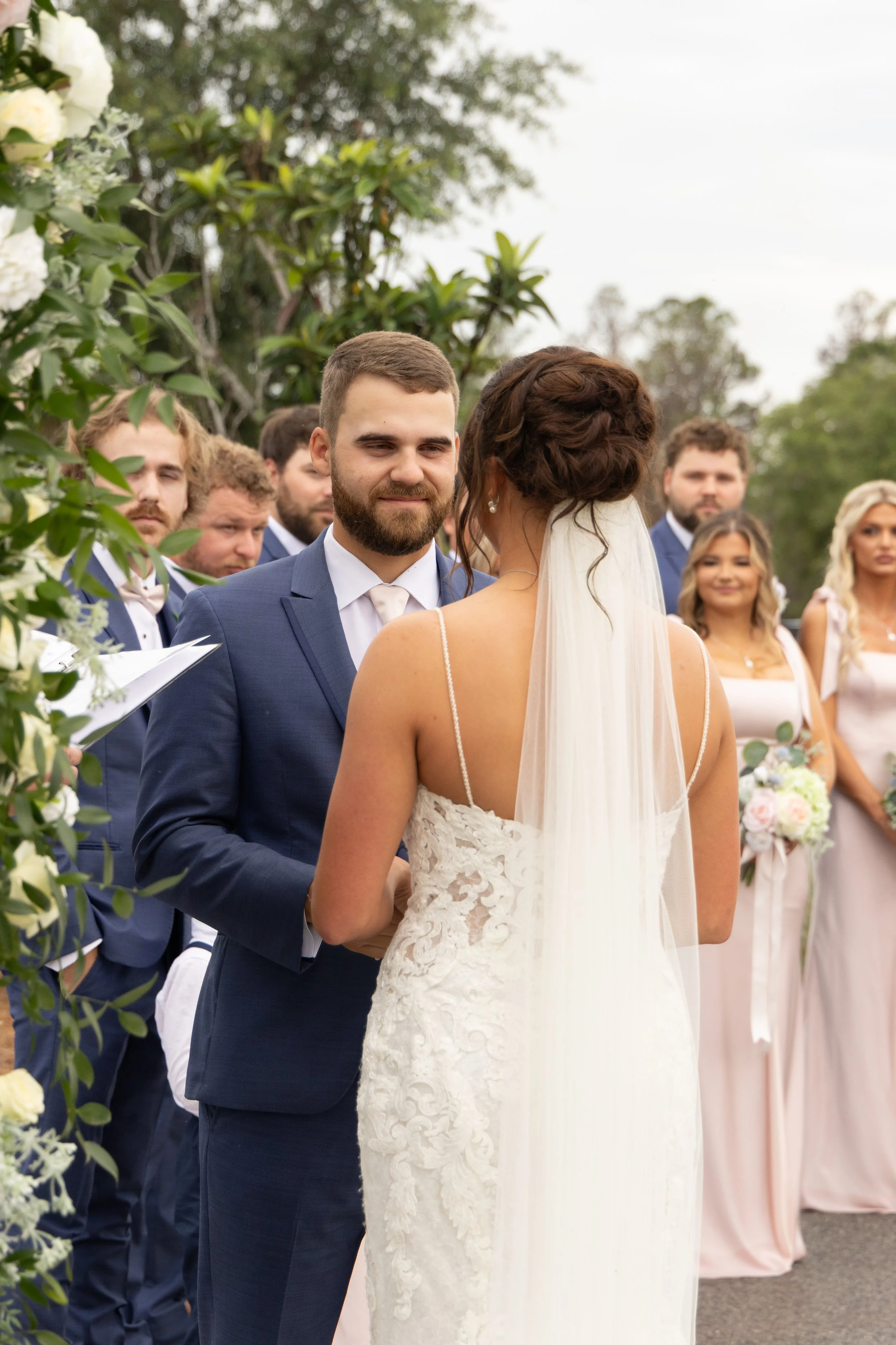 Dylan smiles at Emma as they hold hands during their outdoor wedding ceremony at Red River Estate in Waynesville, Georgia.