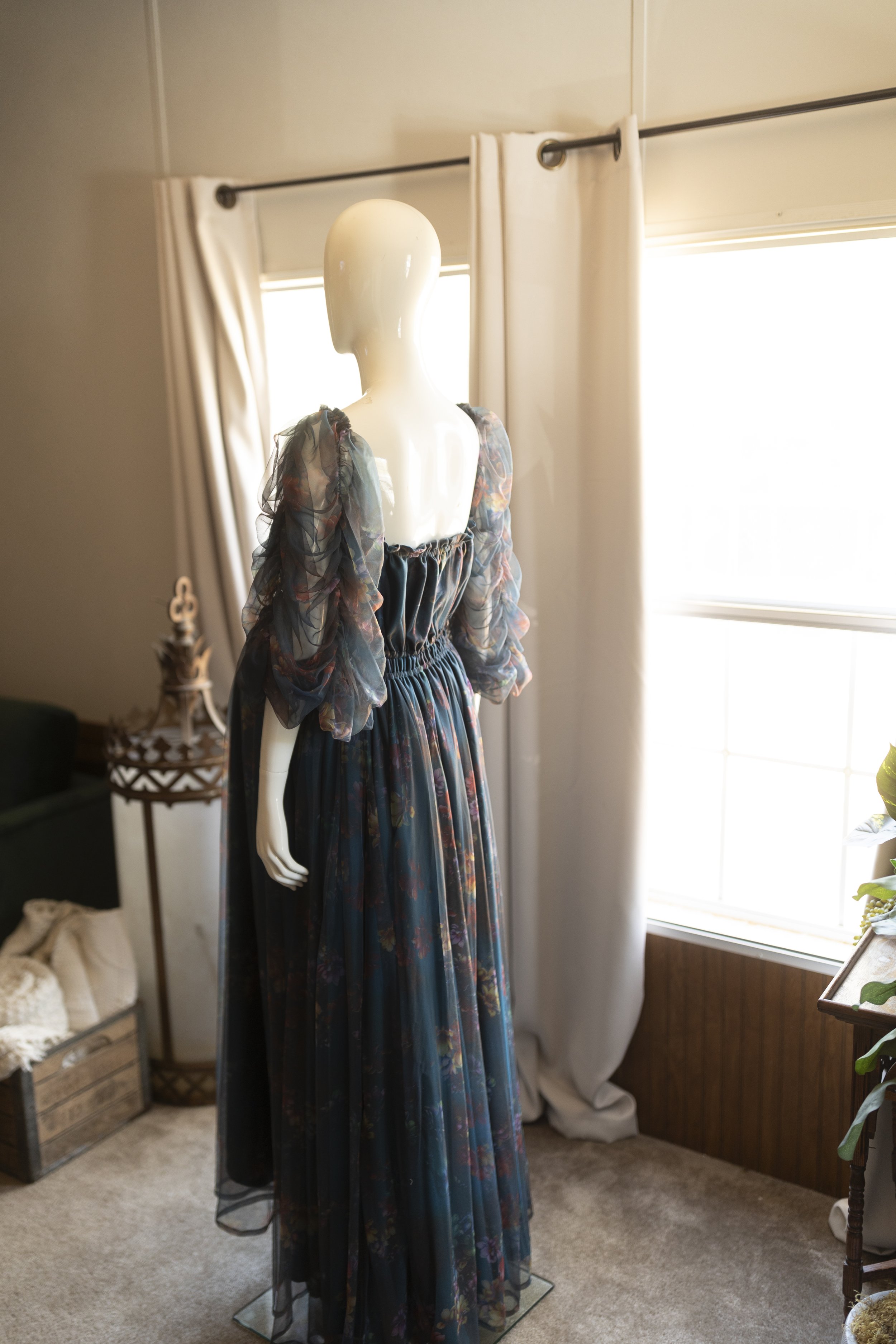 Back view of a dark floral chiffon maternity gown with sheer puff sleeves, a gathered elastic back, and a flowing skirt displayed on a mannequin.