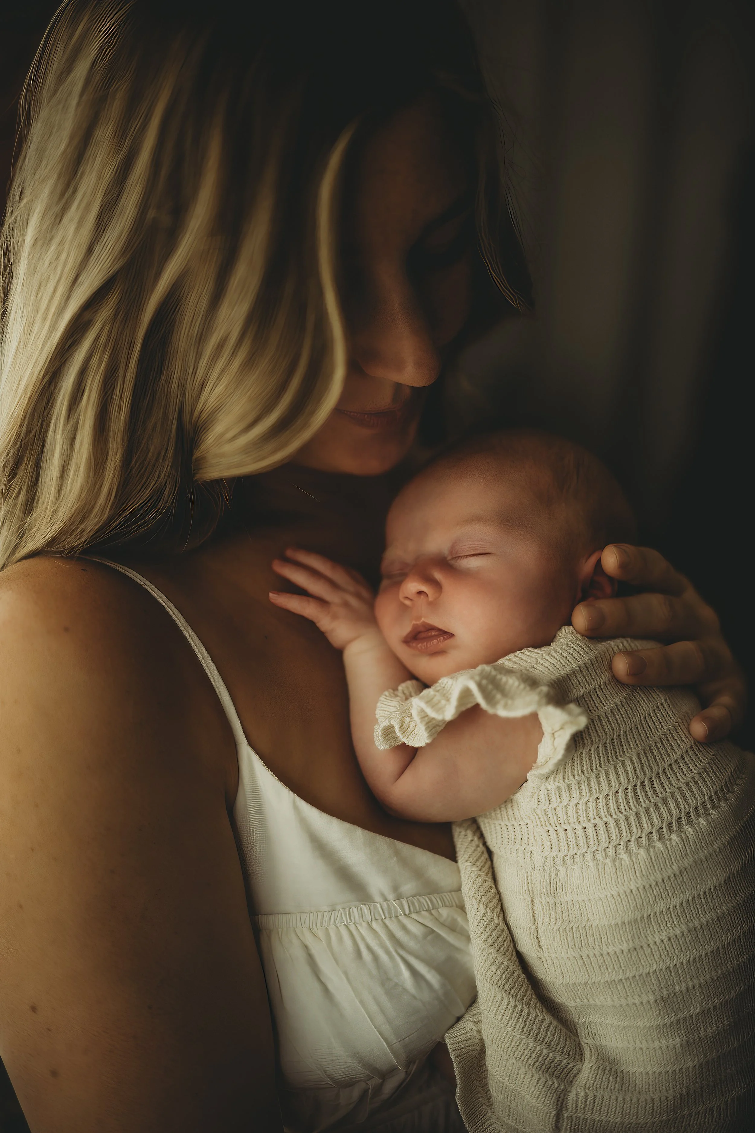 Newborn sleeping peacefully against their mother’s shoulder, wrapped in a textured blanket and held close in warm, intimate light.
