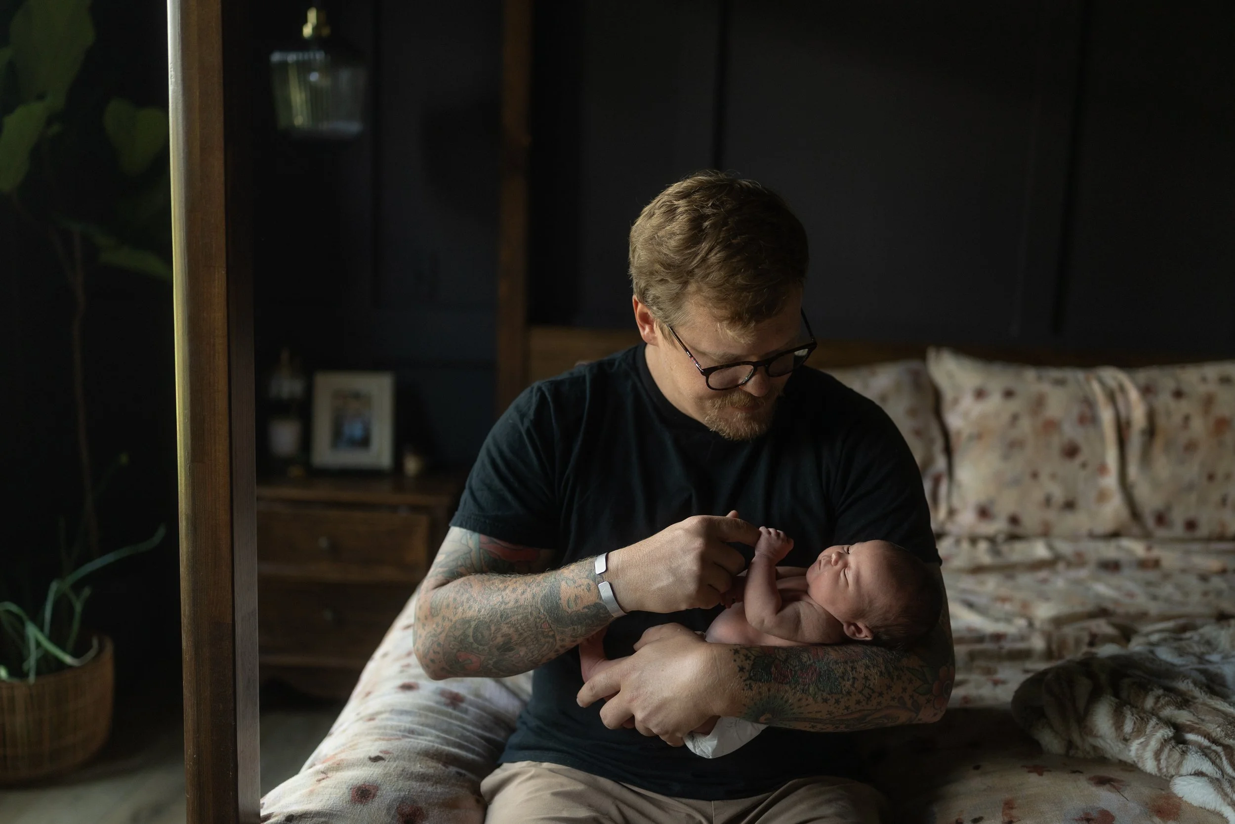 Father sits on the bed holding his newborn in his arms, smiling down as they share a quiet one-on-one moment during an intimate in-home lifestyle newborn photography session.