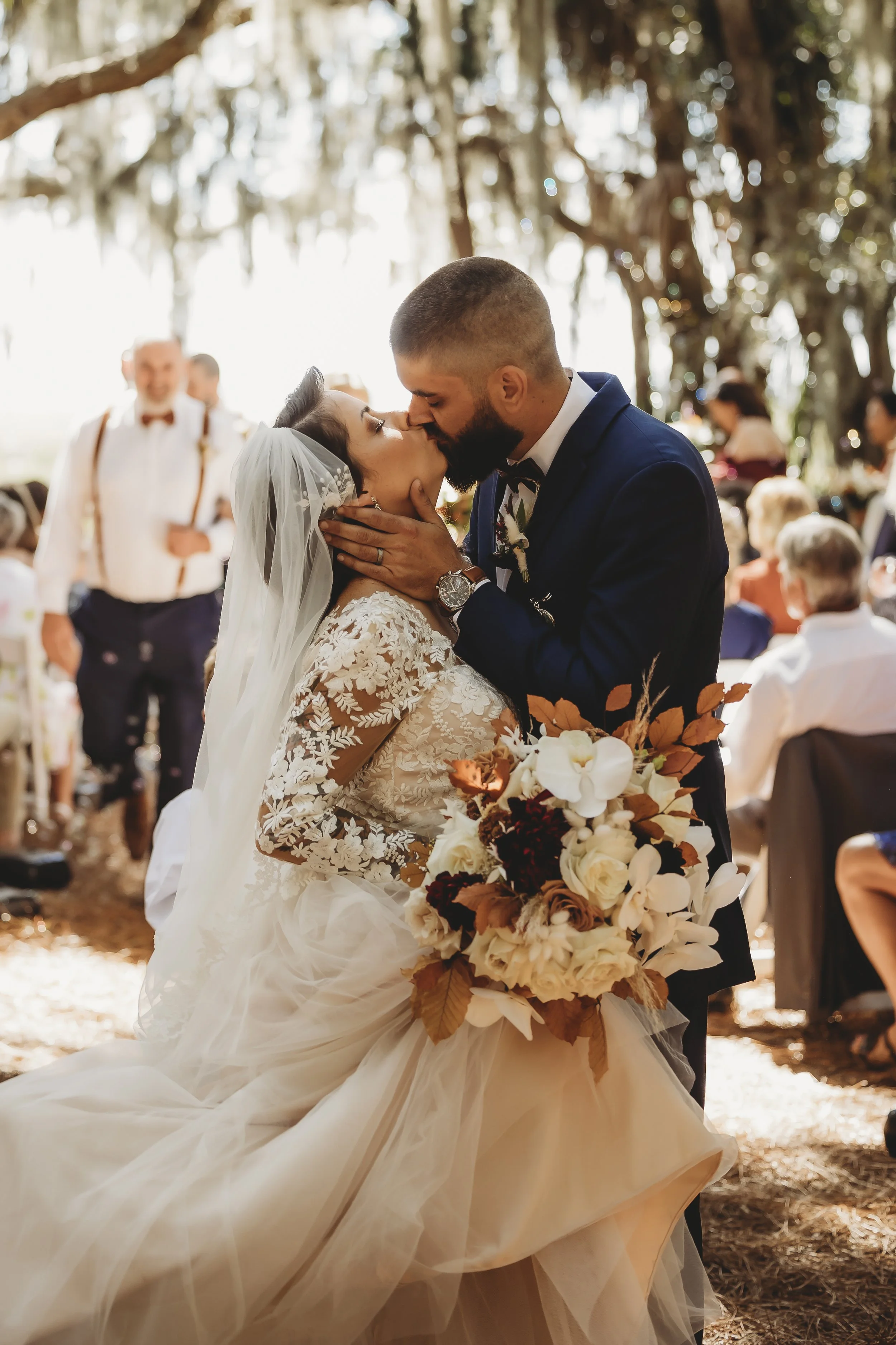 A bride and groom share a kiss at their outdoor wedding ceremony, with the bride holding a bouquet of flowers and wearing a lace wedding dress, and the groom in a navy suit.