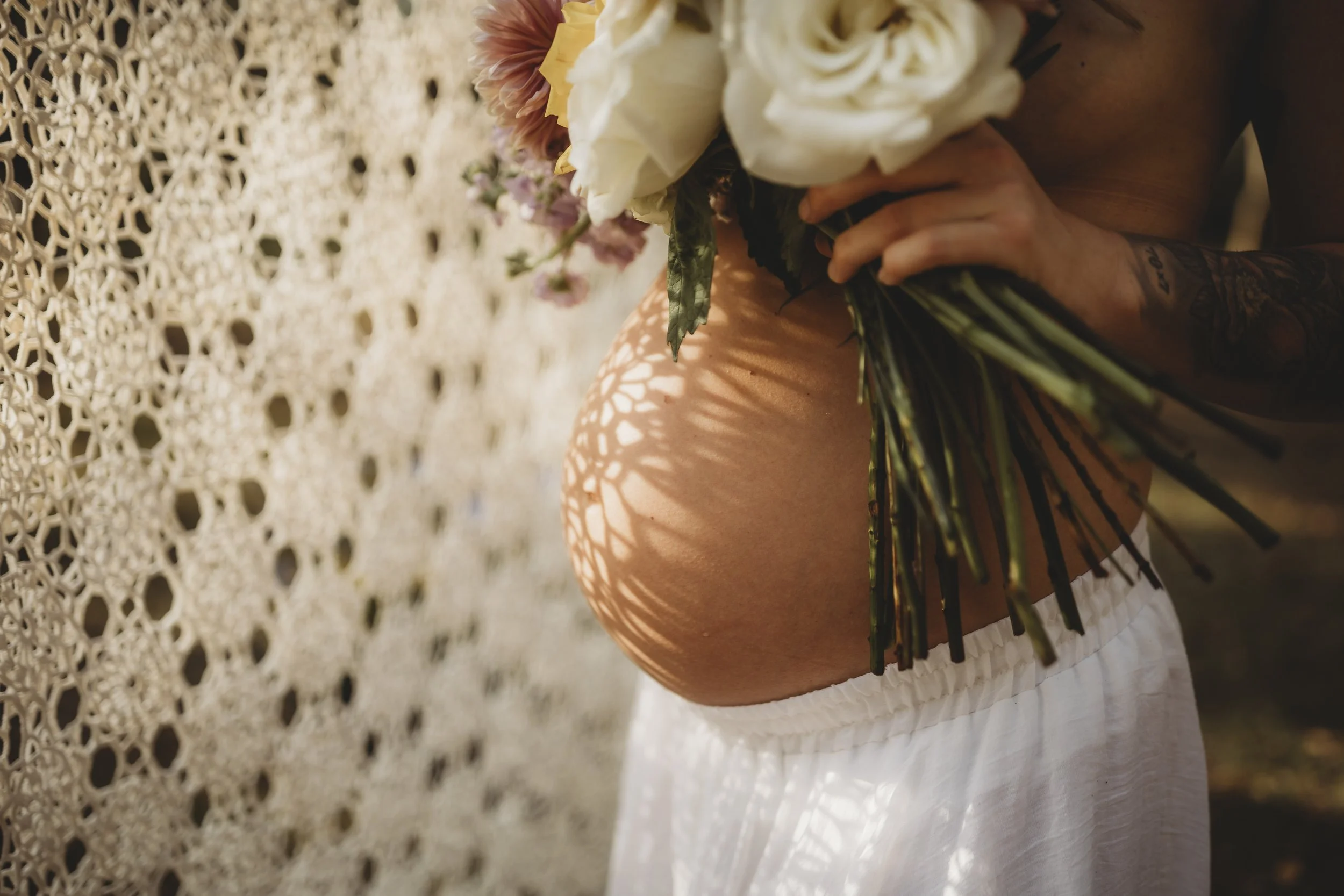 Warm sunlight casts delicate shadows across a pregnant belly as a mother holds a bouquet close, honoring the beauty of new life