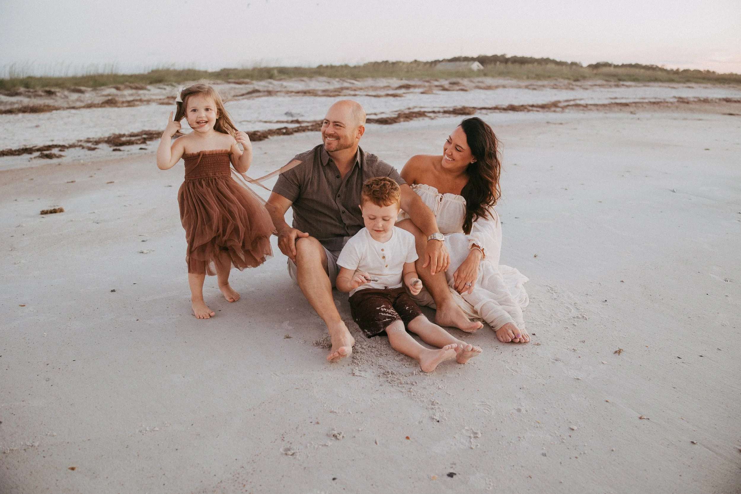 A family of four sitting and standing on a sandy beach with grass dunes and a cloudy sky in the background, smiling and enjoying their time together.