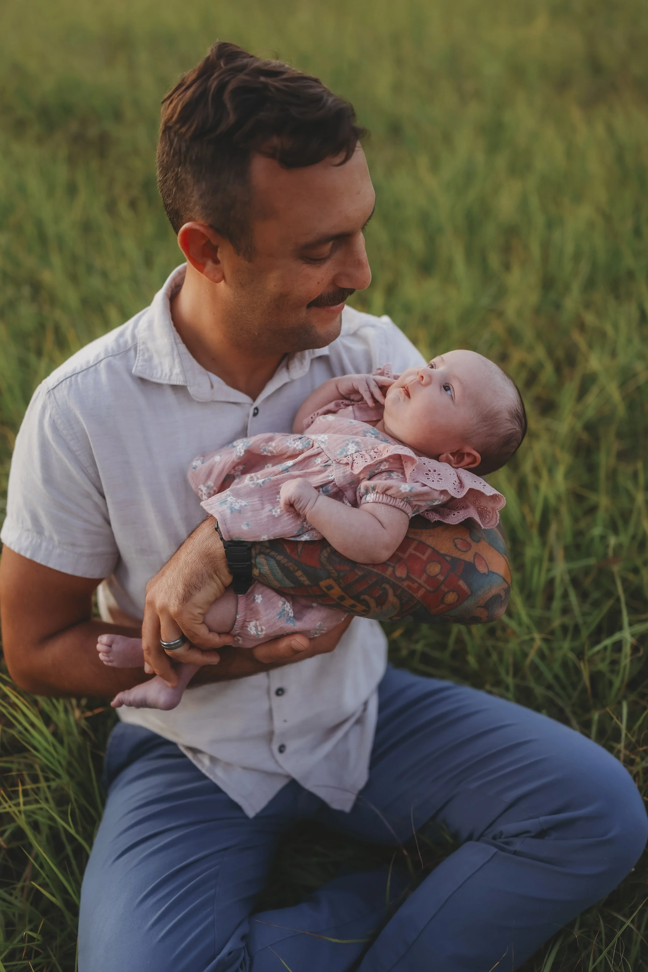 A father sits in a grassy field at sunset, smiling down at his newborn daughter as he gently cradles her in his arms during a heartfelt family session.