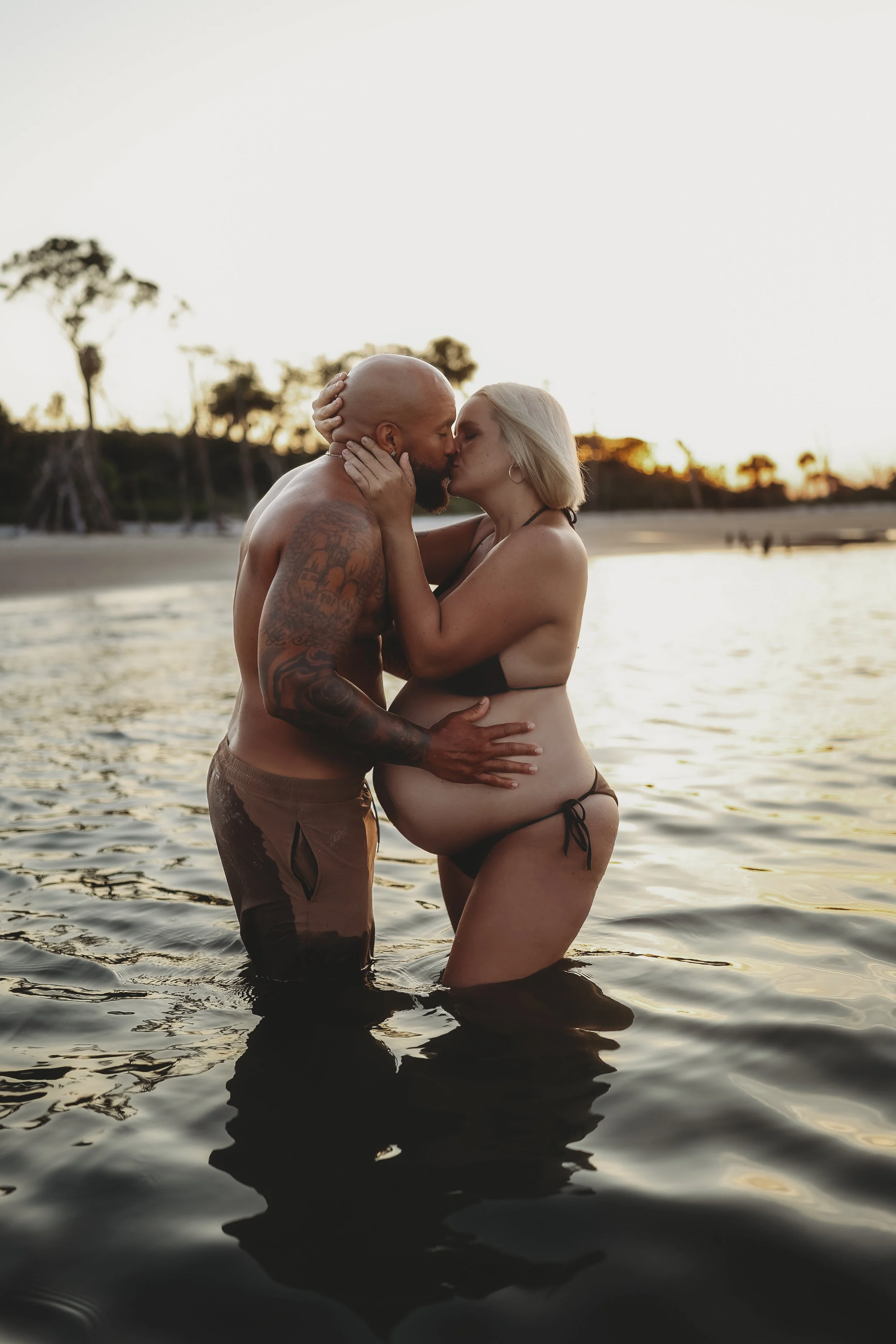 Beach maternity portrait of an expecting couple kissing while standing together in shallow water at sunset.