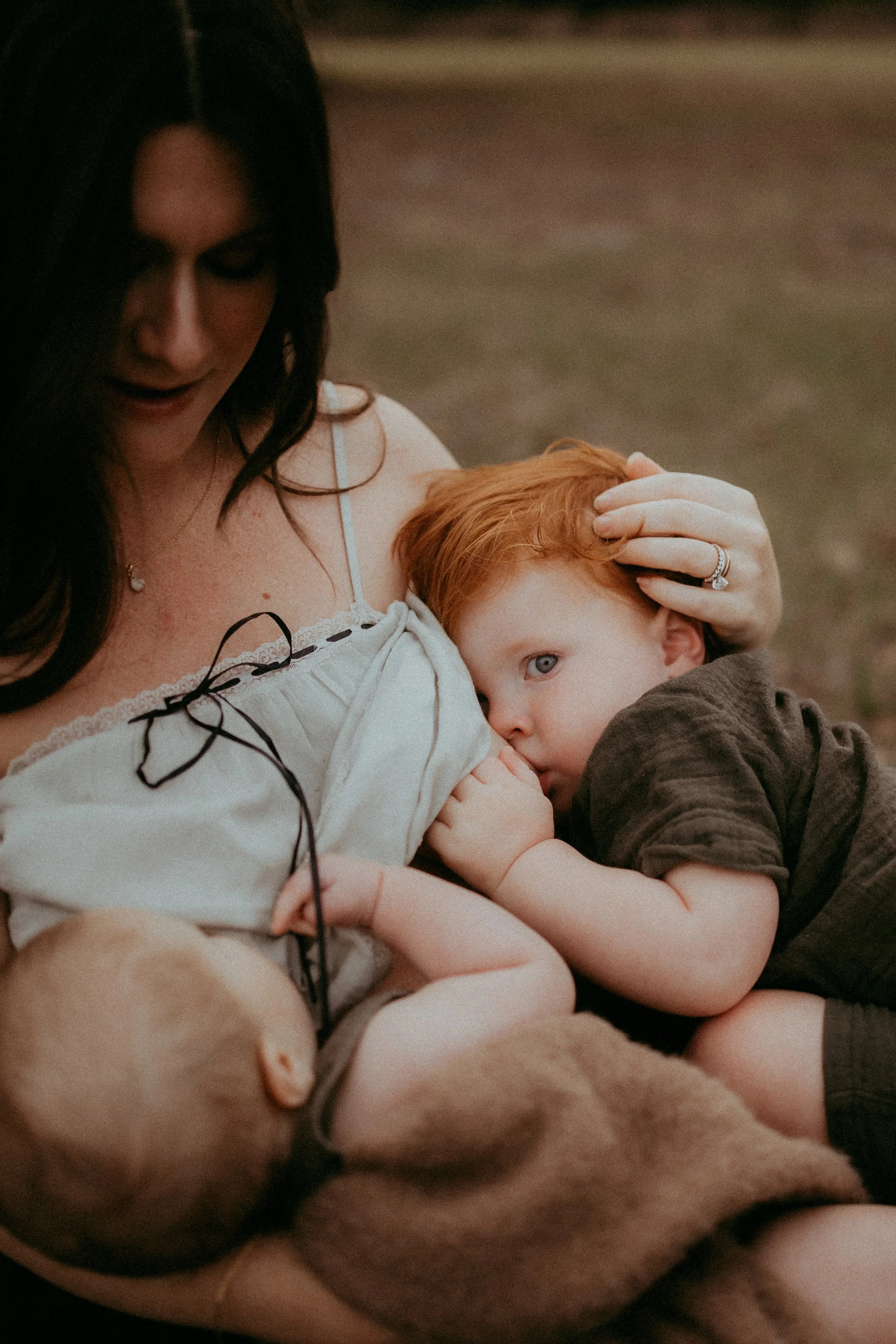 A mother tandem breastfeeds her older child and baby while they share a quiet moment together in an open field.