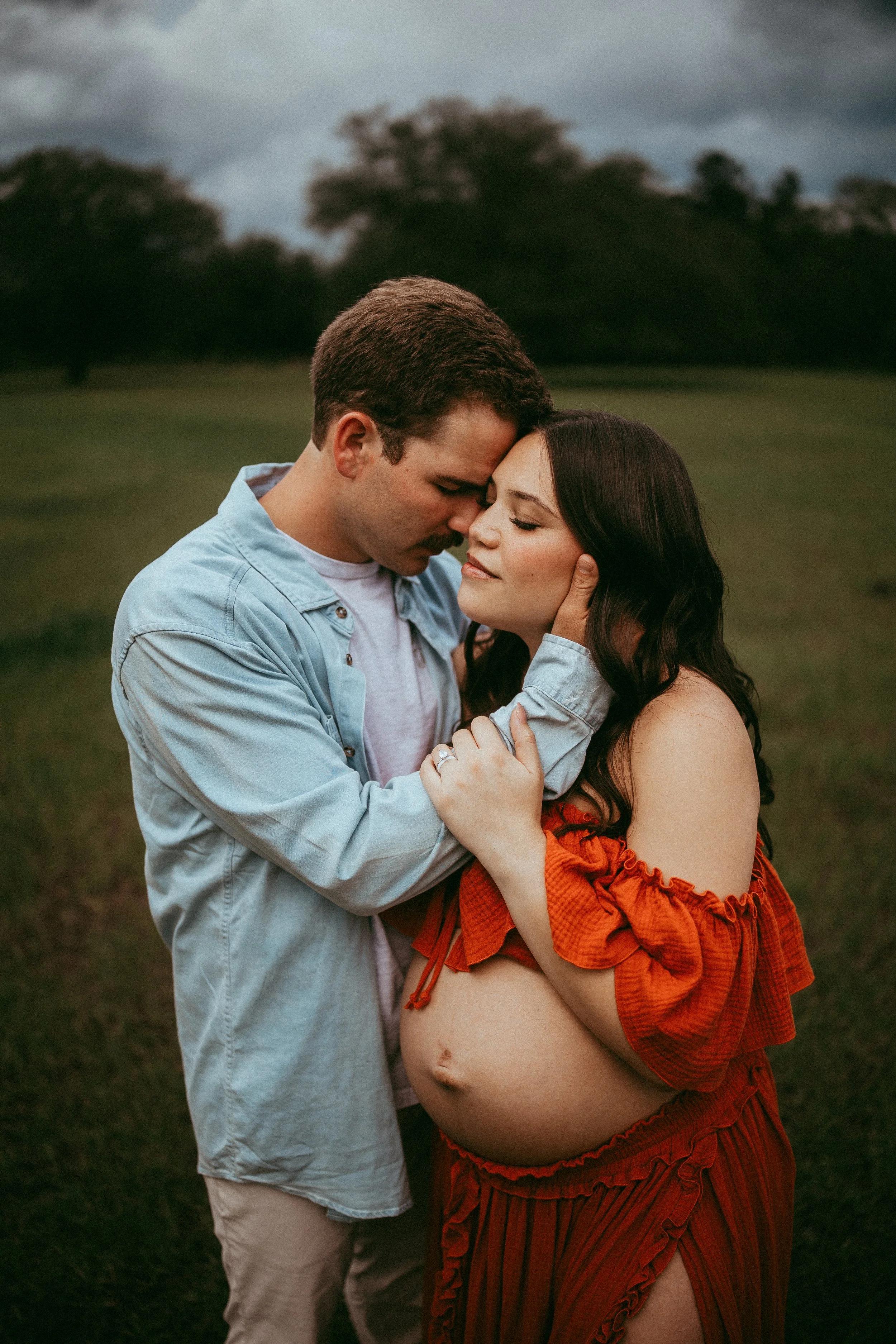 A man and pregnant woman embrace outdoors on cloudy day, with trees in background.