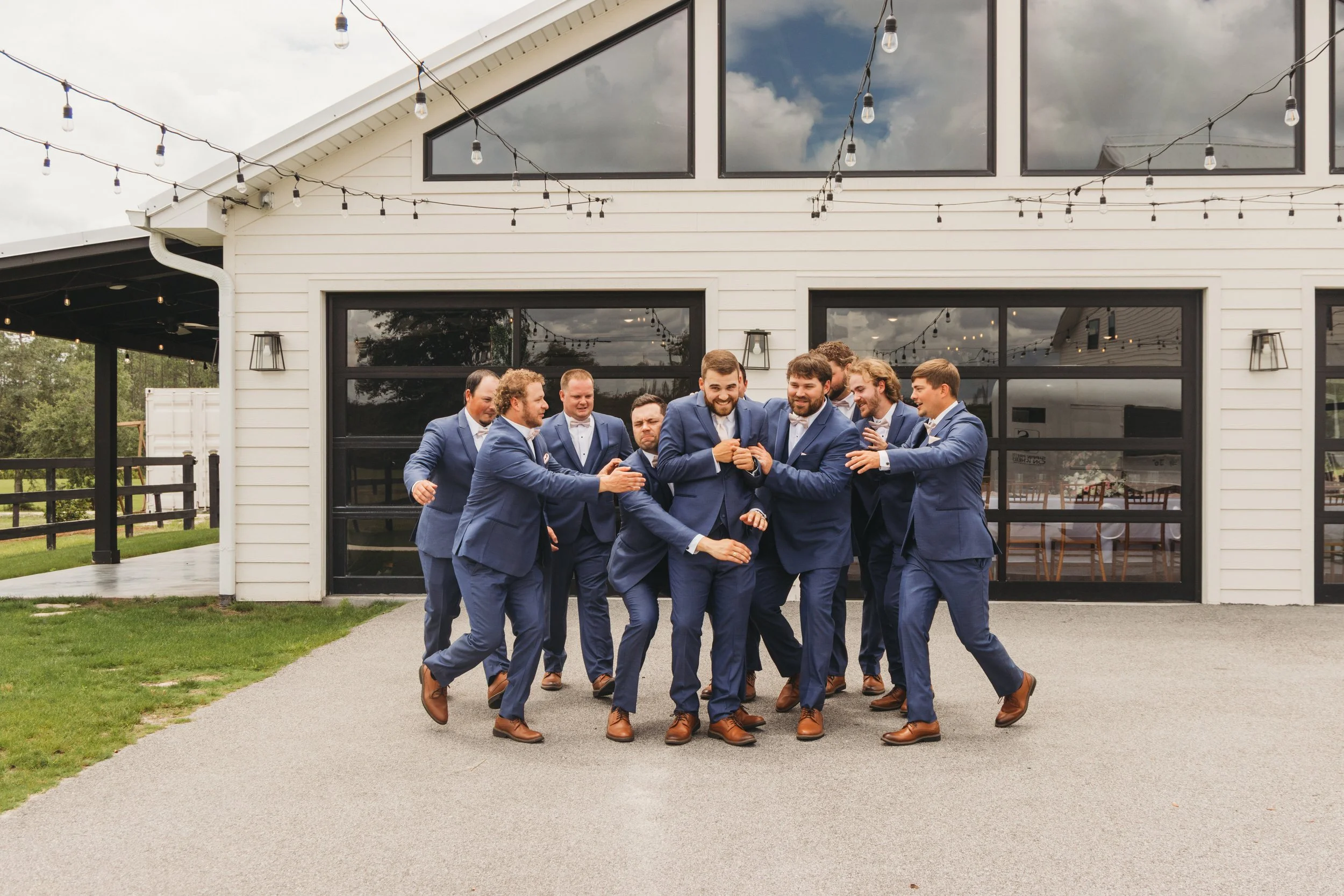 Dylan laughs as his groomsmen playfully crowd around him outside the white reception barn at Red River Estate.