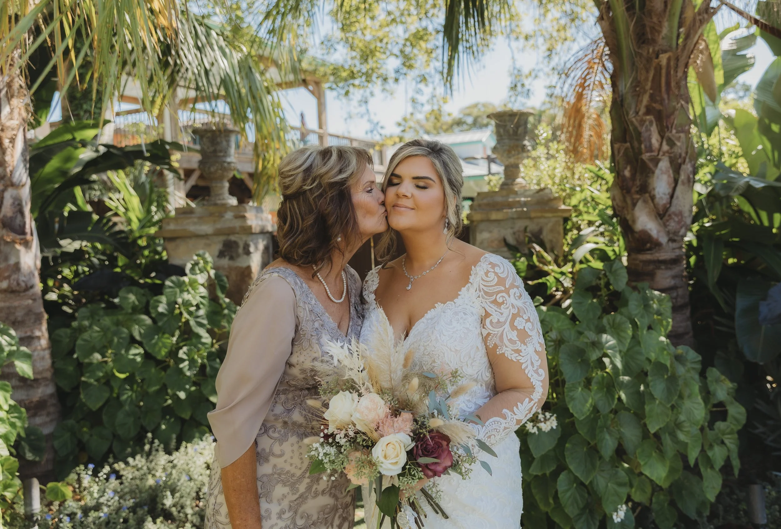Mother kissing the bride on the cheek while she holds her bouquet in a garden