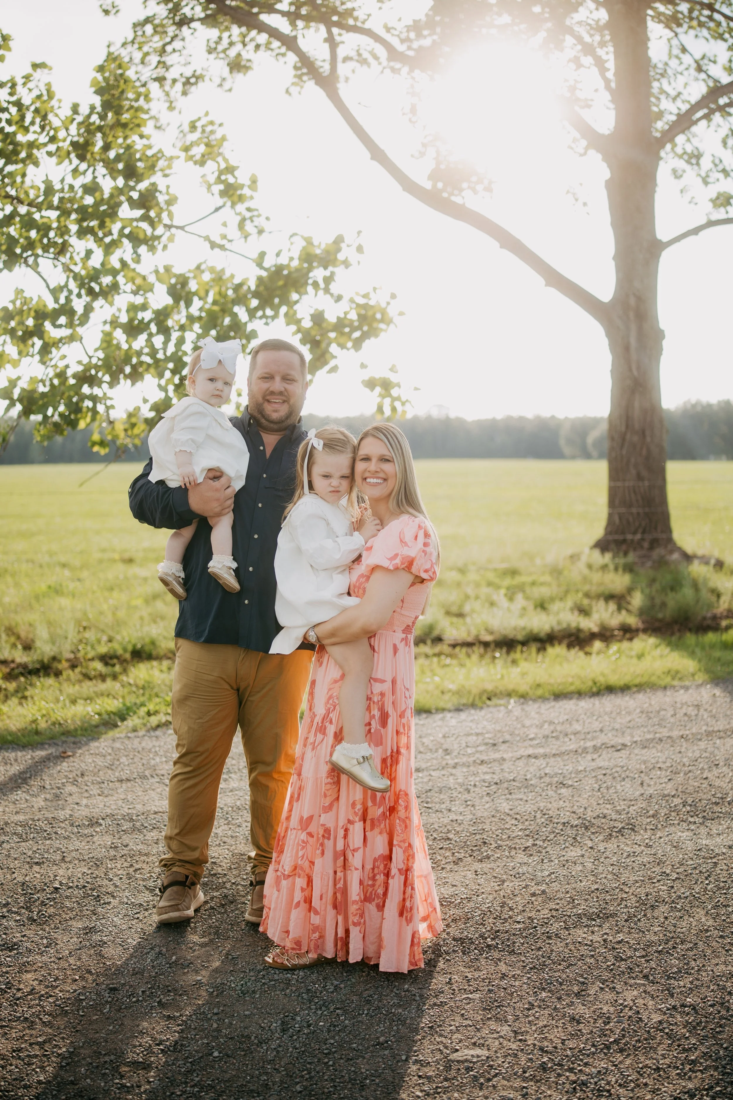 A family of four stands together beneath sunlit oak trees, each parent holding a daughter as they share a joyful outdoor family portrait filled with love, warmth, and timeless connection.