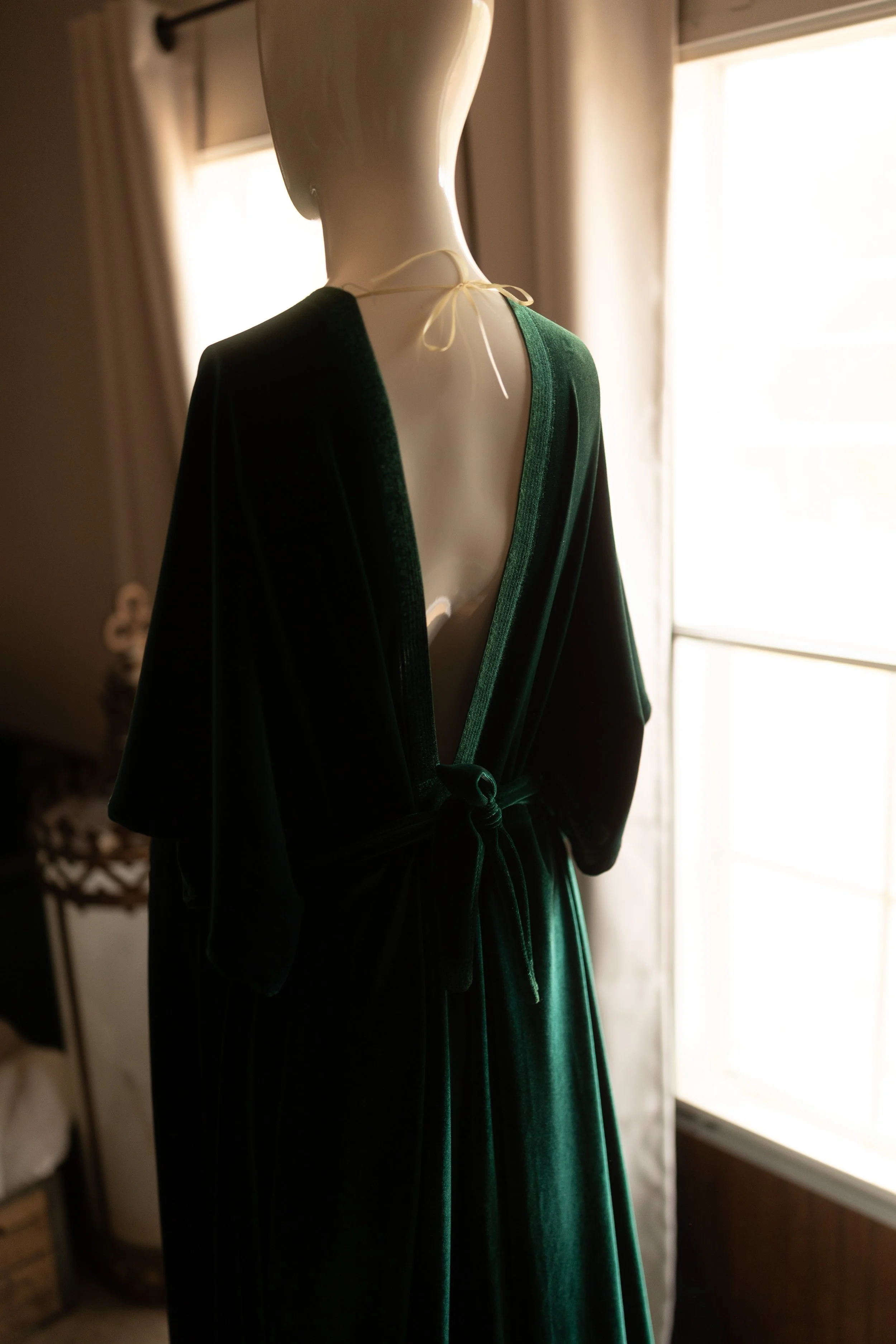 Deep emerald velvet maxi dress with a wrap-style V-neck bodice, open back with tie detail, long sleeves, and a flowing skirt, styled on a mannequin in soft natural light.
