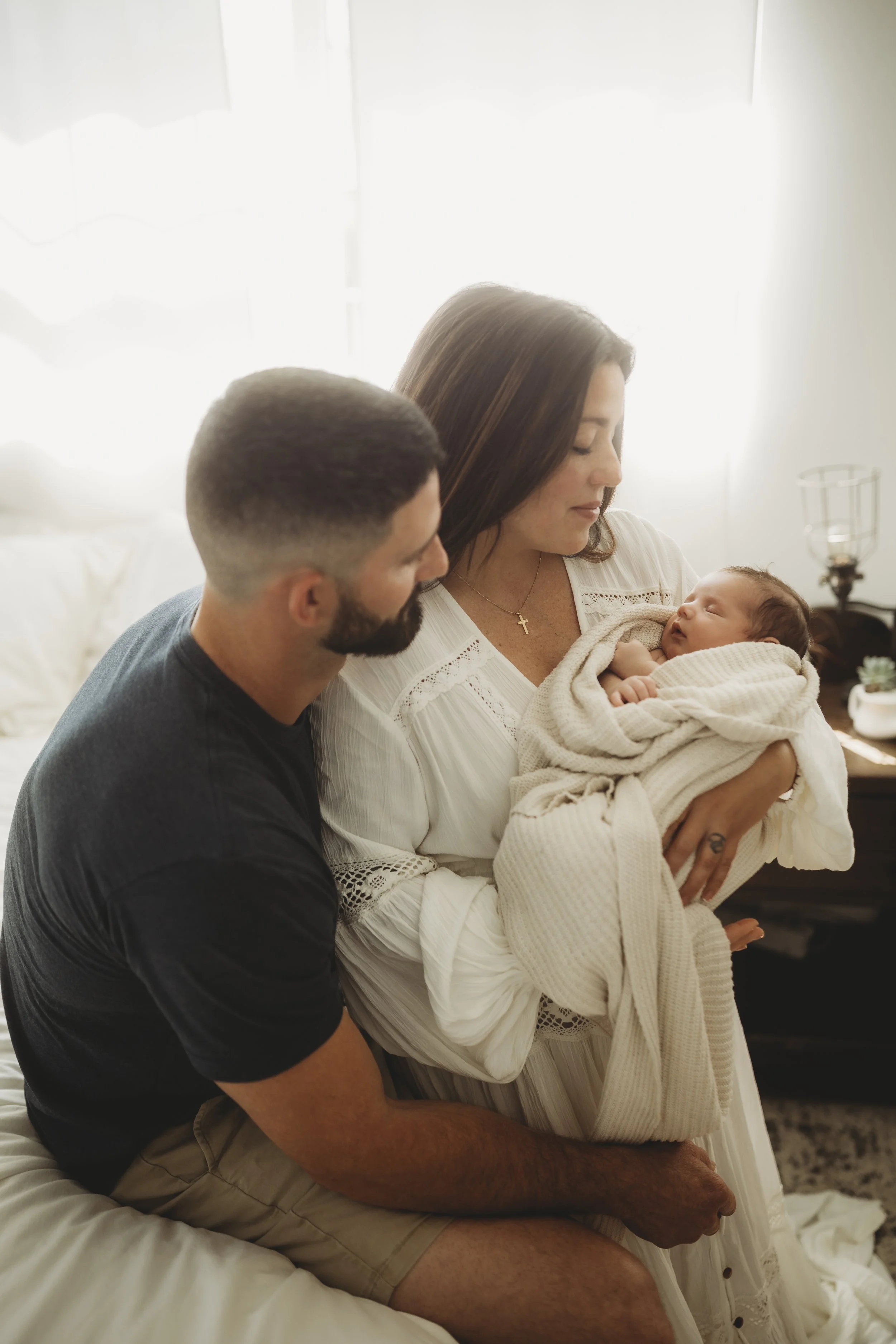 Parents seated together while a mother holds her newborn, wrapped gently in soft fabric and natural light.