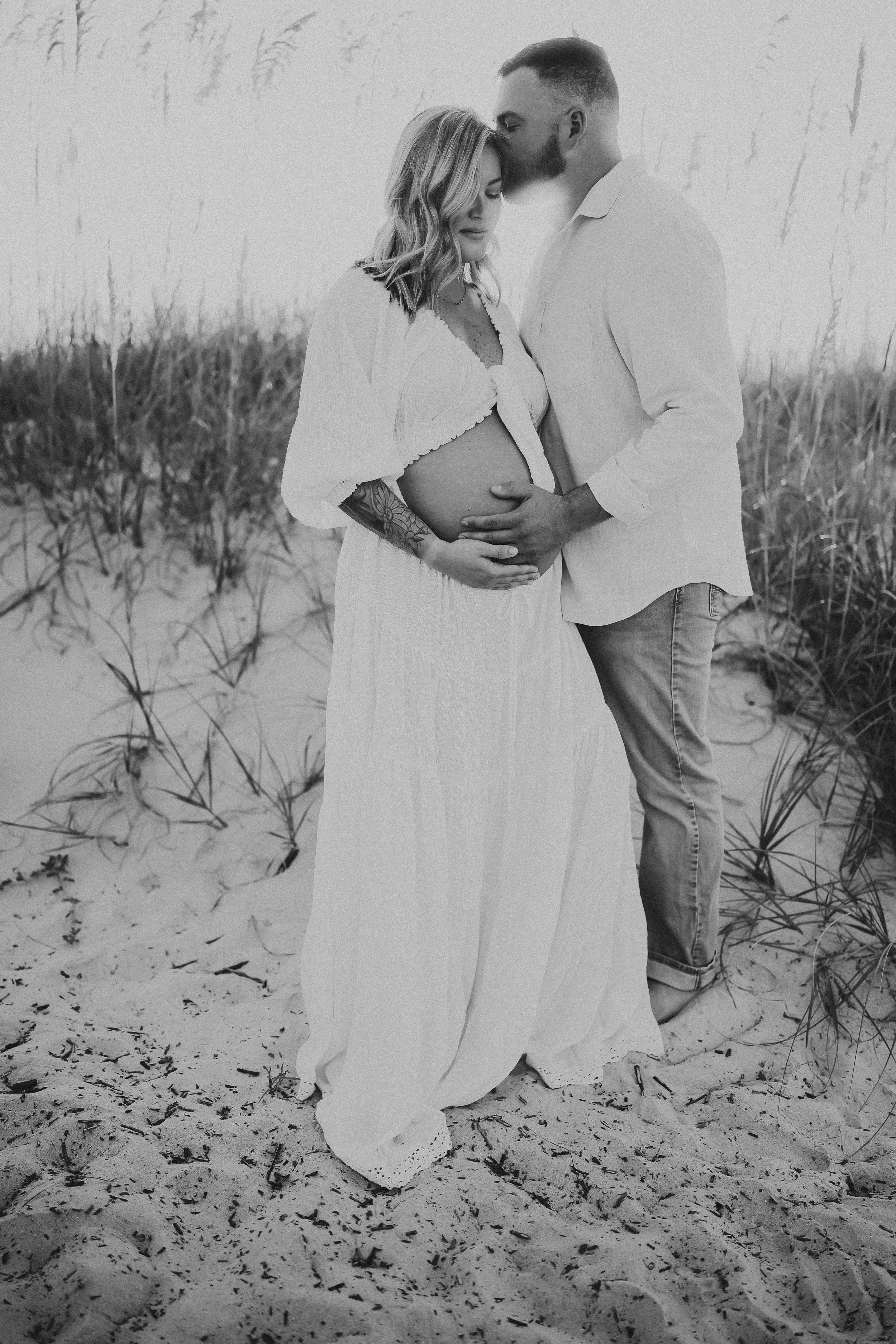 Black and white maternity portrait of an expectant couple standing in tall beach grass, with the father gently kissing the mother’s forehead as they cradle her baby bump.