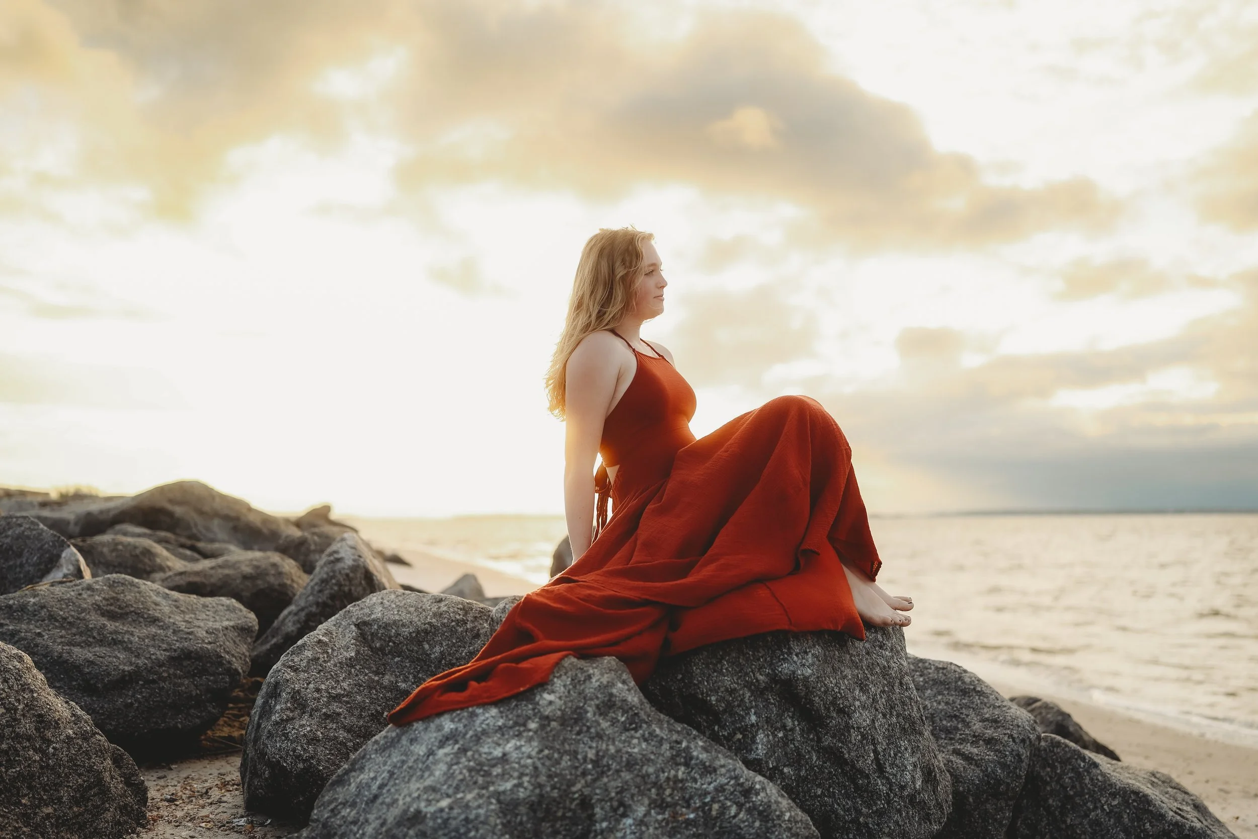 A teenage daughter sits gracefully on the shoreline rocks in a flowing rust-colored dress at sunset.