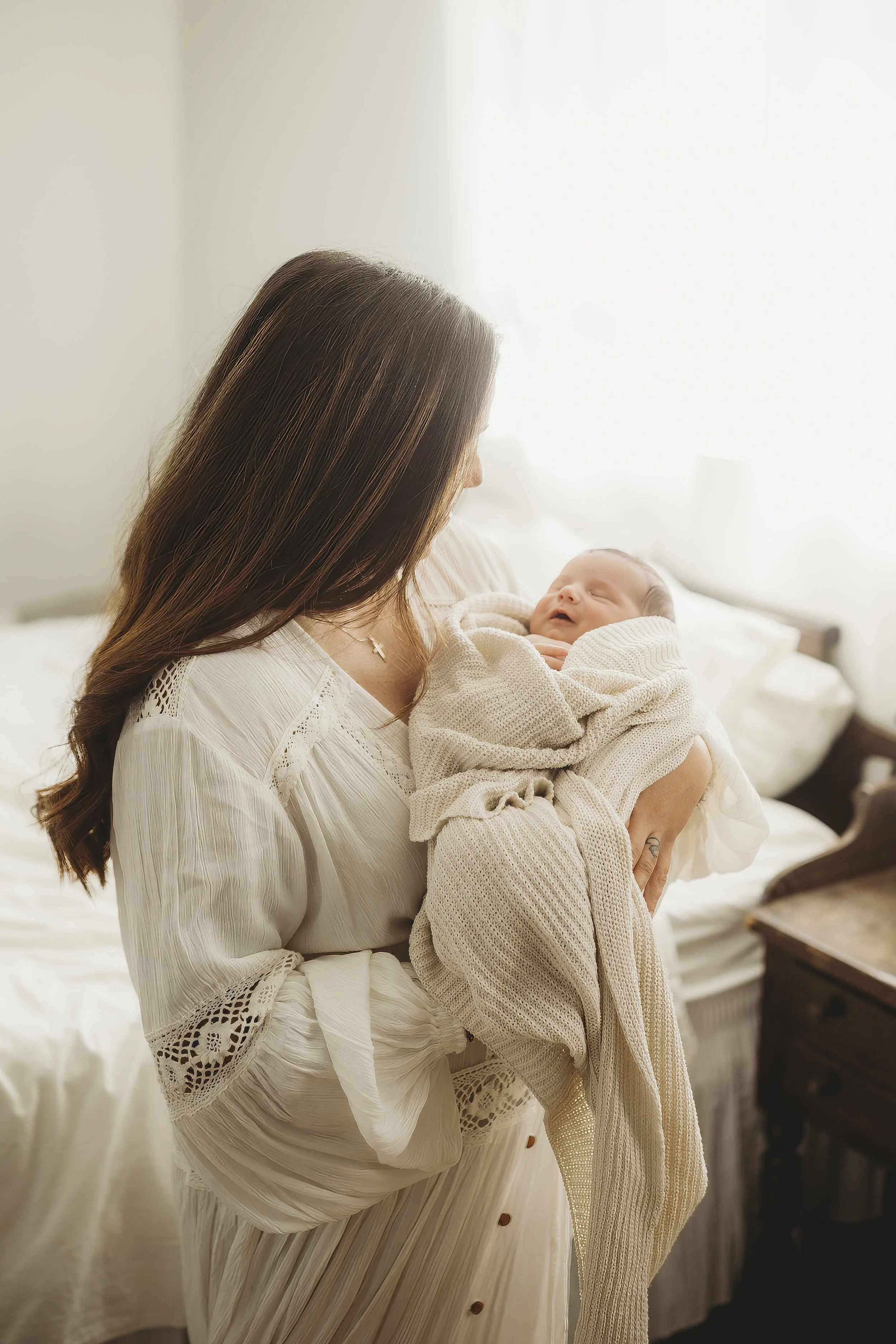 Mother holding newborn in soft window light, wearing ivory lace boho dress styled for both maternity and postpartum portraits.