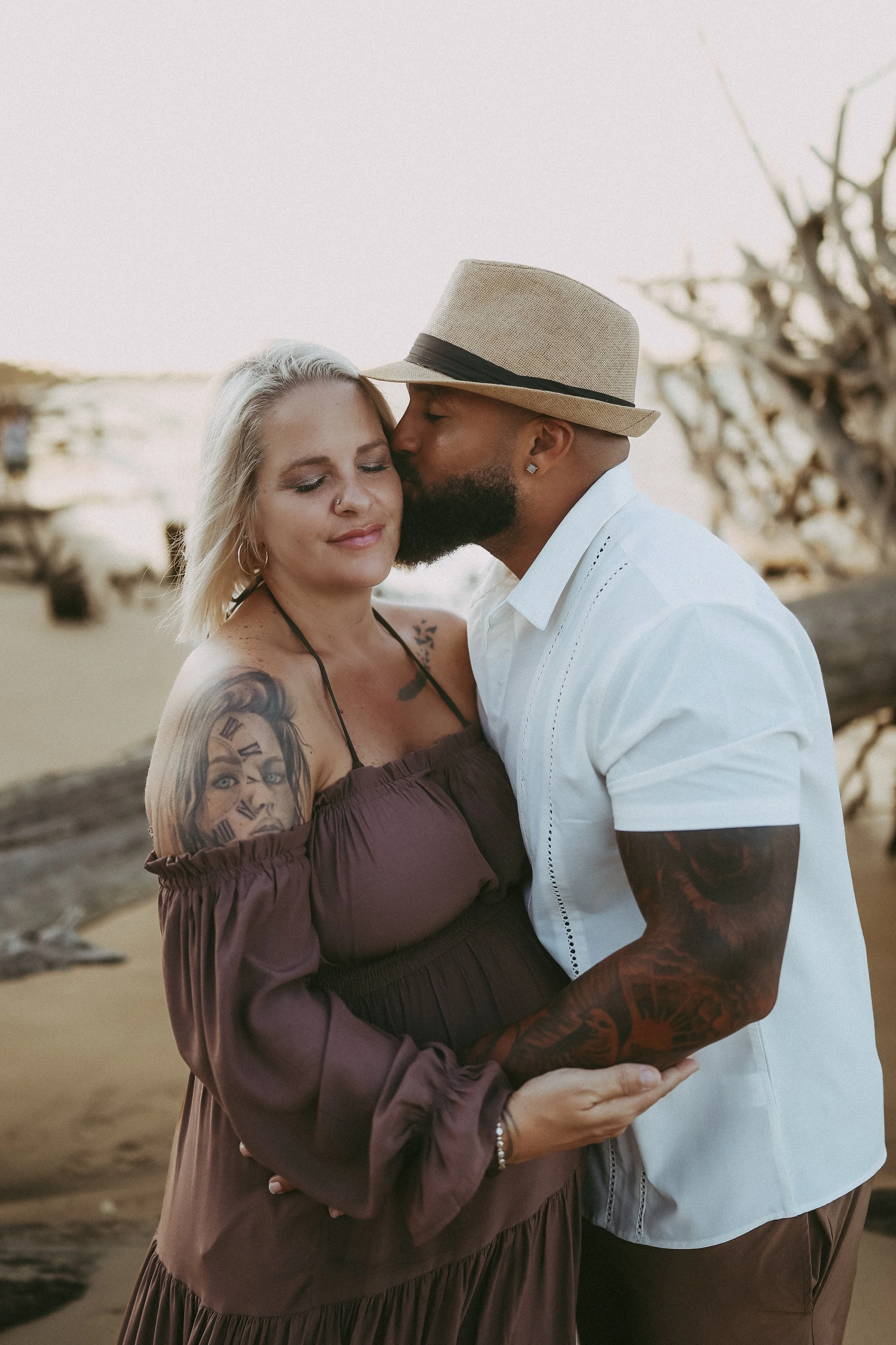 Romantic maternity photo of an expectant couple standing on the beach, sharing a quiet embrace as the partner kisses the mother’s temple during sunset.