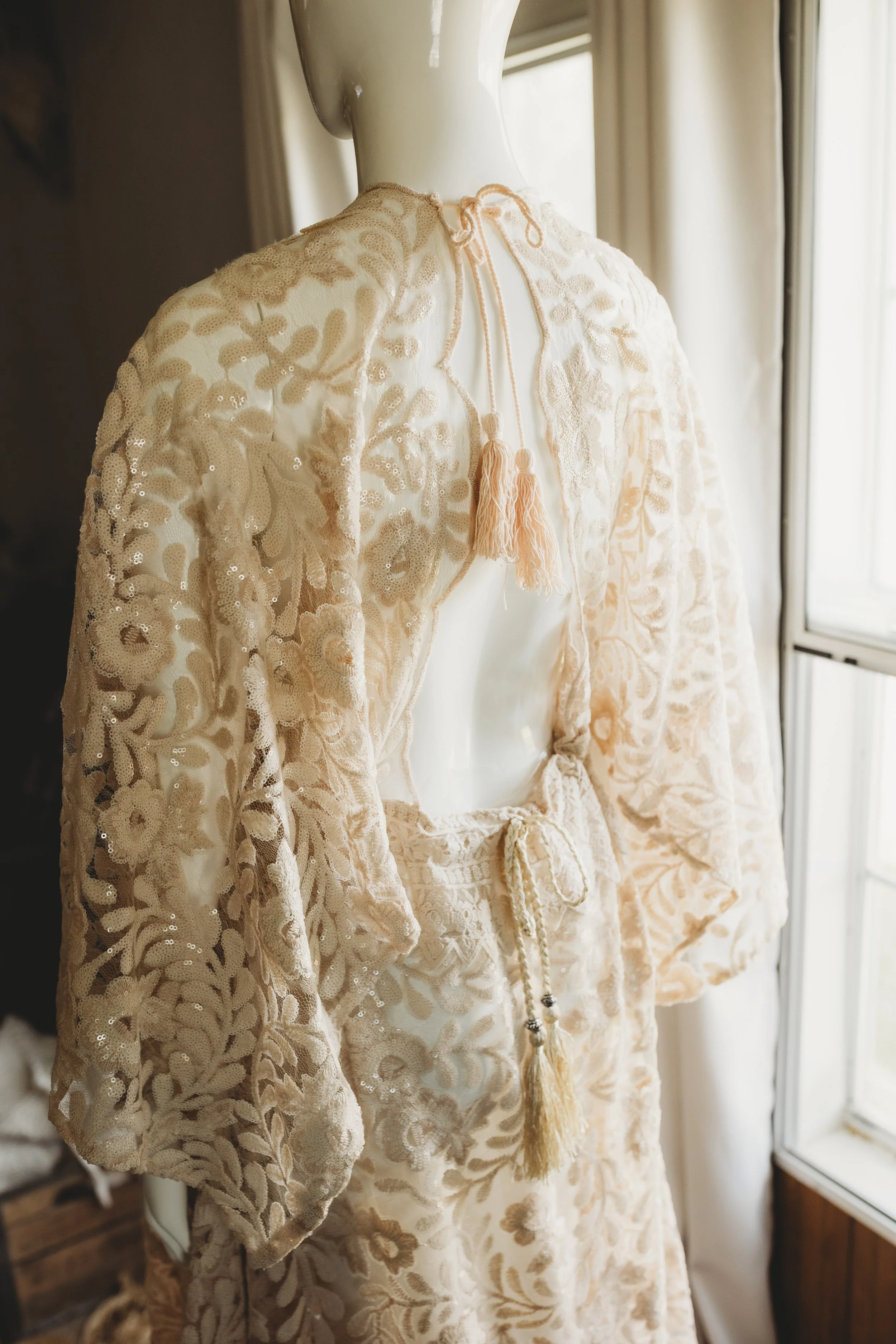 Ivory lace gown with long sleeves, an open back, and delicate tassel ties, styled for maternity and portrait sessions.