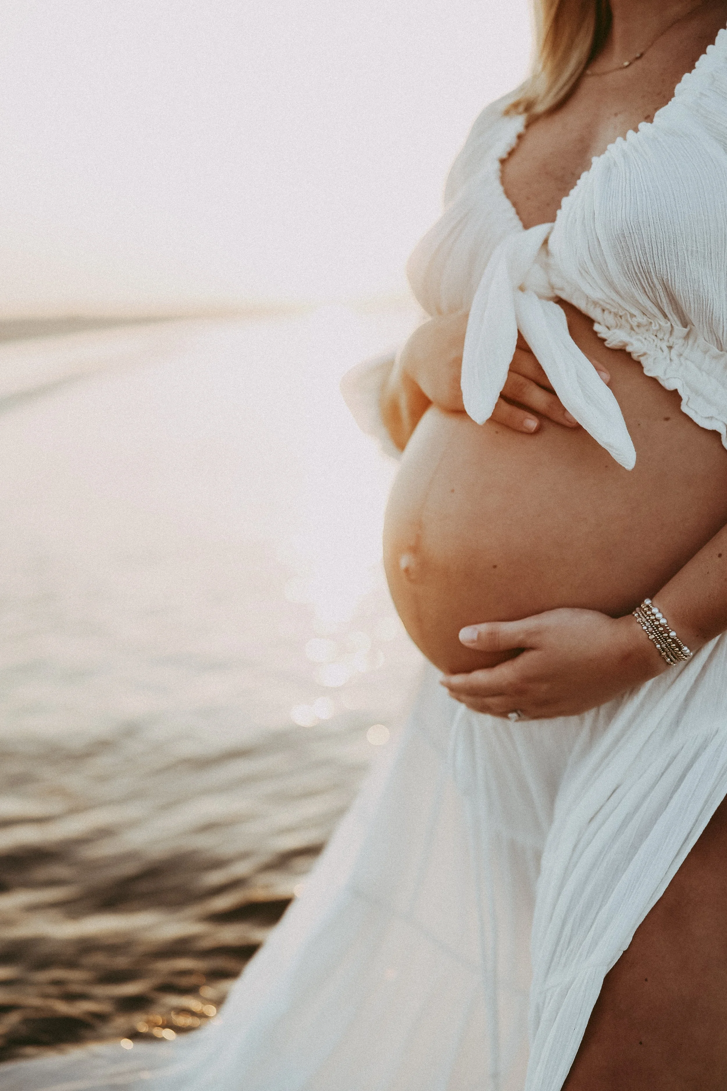 Close-up maternity photo of an expectant mother’s baby bump at sunset, her hands gently cradling her belly as soft light reflects off the ocean behind her.