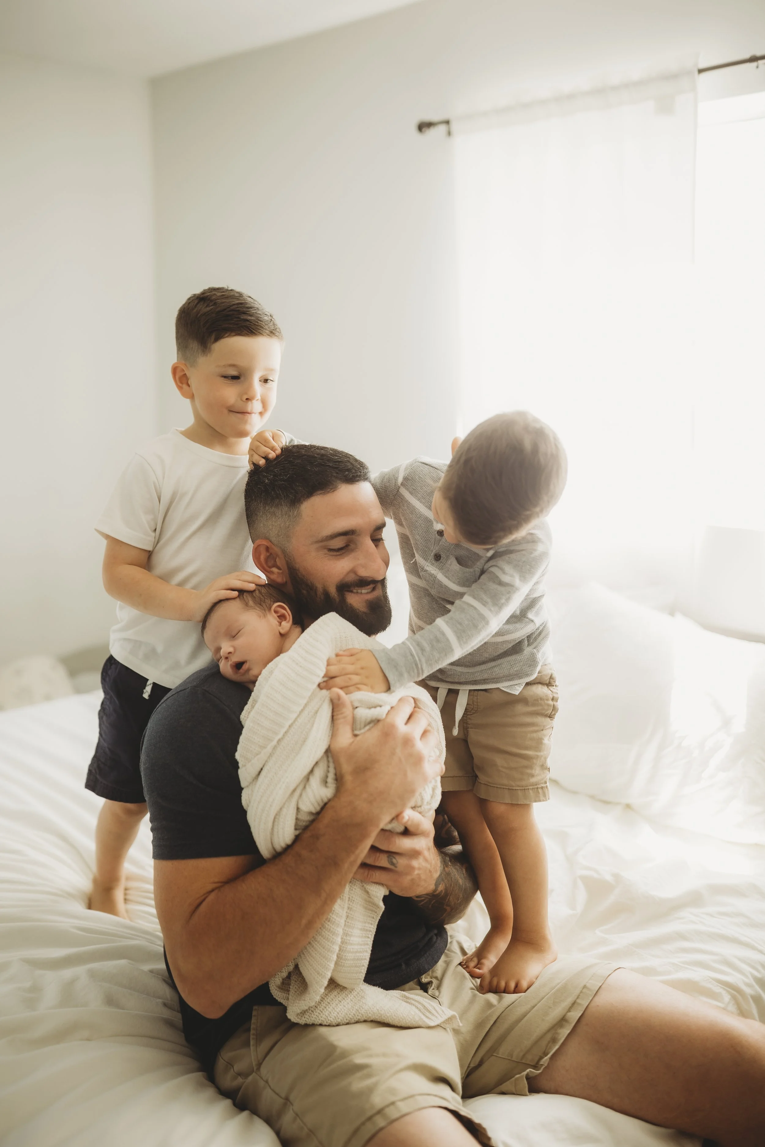 Father sitting on a bed holding his newborn while siblings stand close beside him in soft daylight.