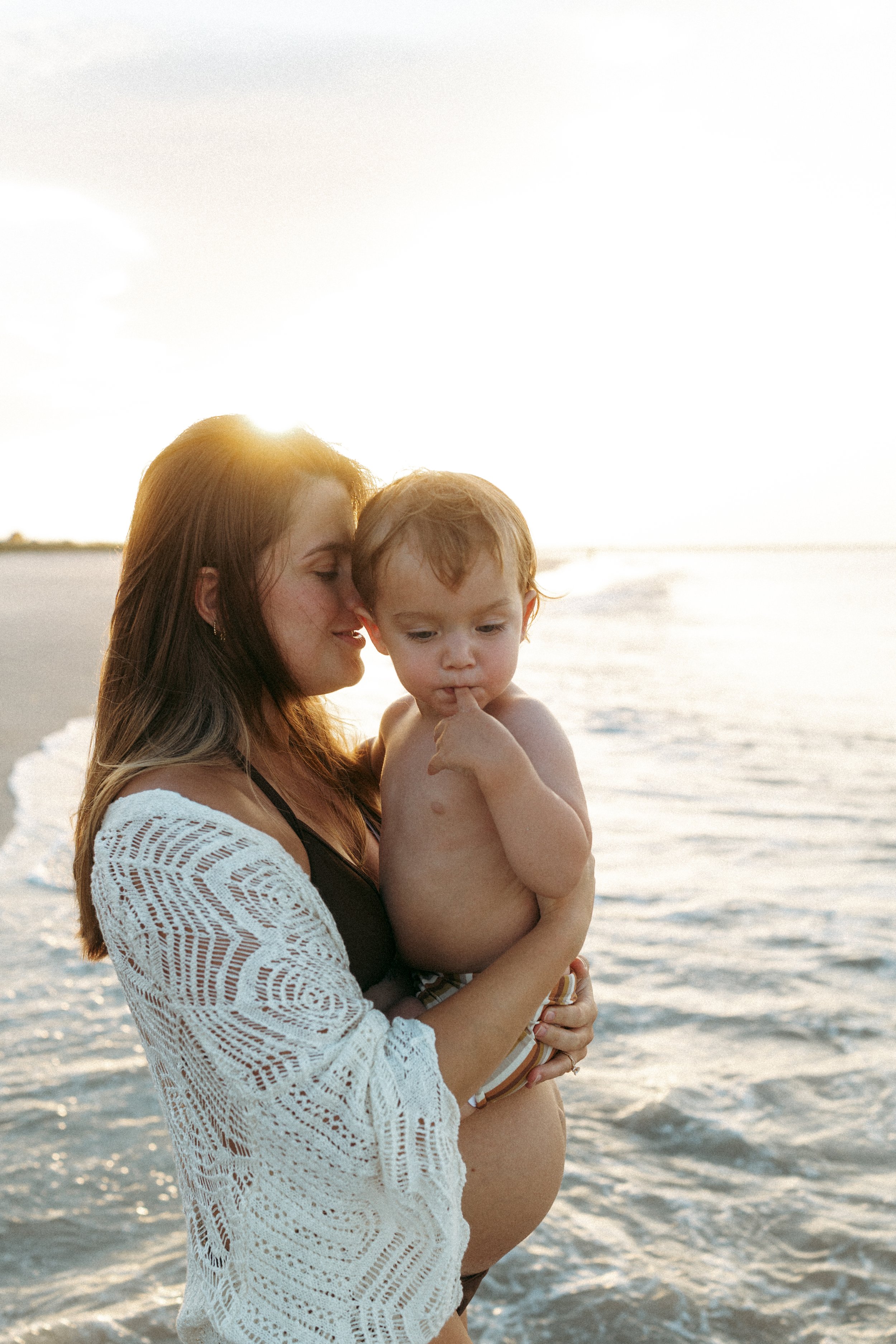 Pregnant mother kisses her toddler's forehead while holding them in gentle ocean waves at sunset on Amelia Island Beach, sharing a quiet moment before welcoming a new baby.