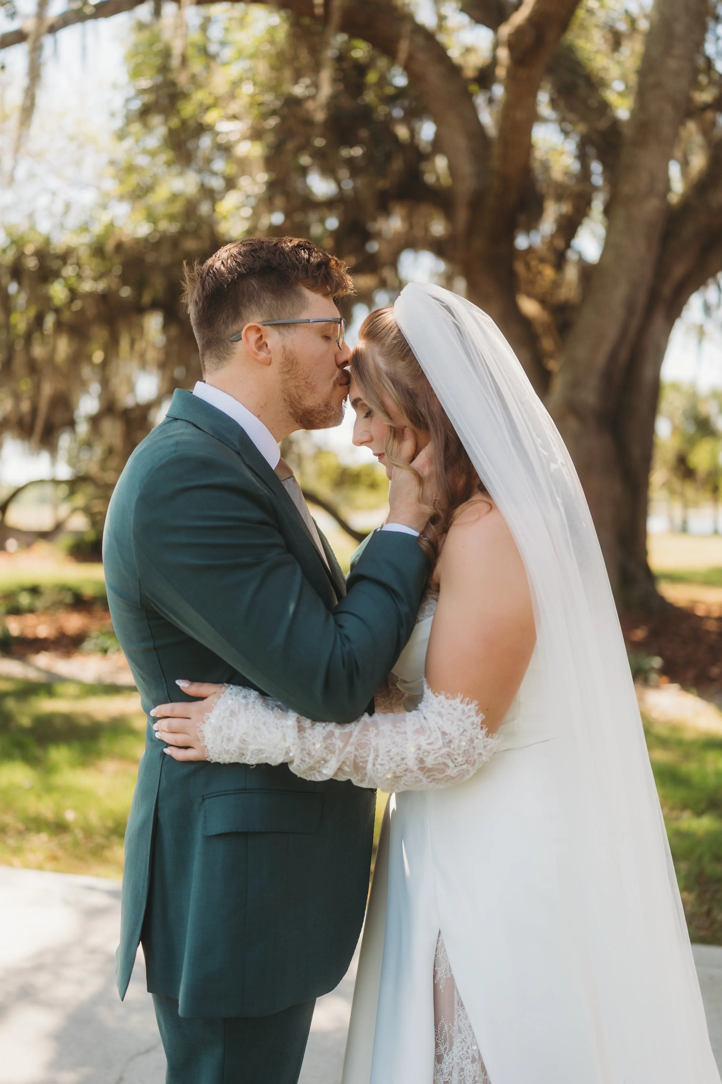 The groom gently kisses the bride's forehead beneath moss-covered oak trees after their ceremony.