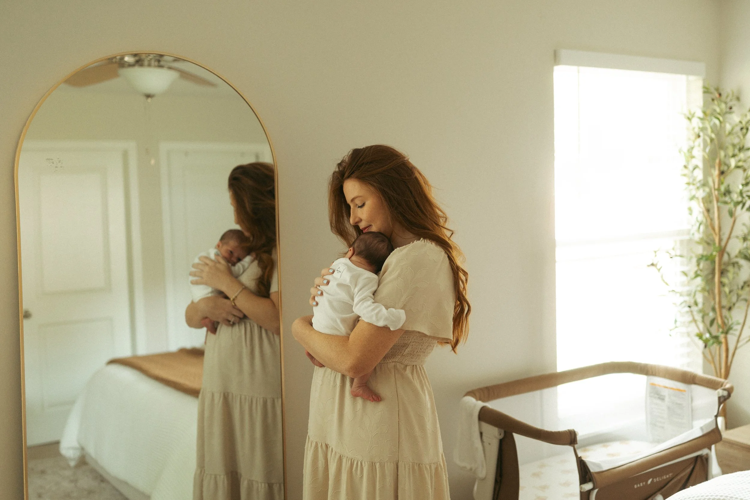 Mother stands in a softly lit nursery, gently holding her newborn against her shoulder as their reflection appears in a full-length mirror, capturing a peaceful moment during an in-home lifestyle newborn photography session.