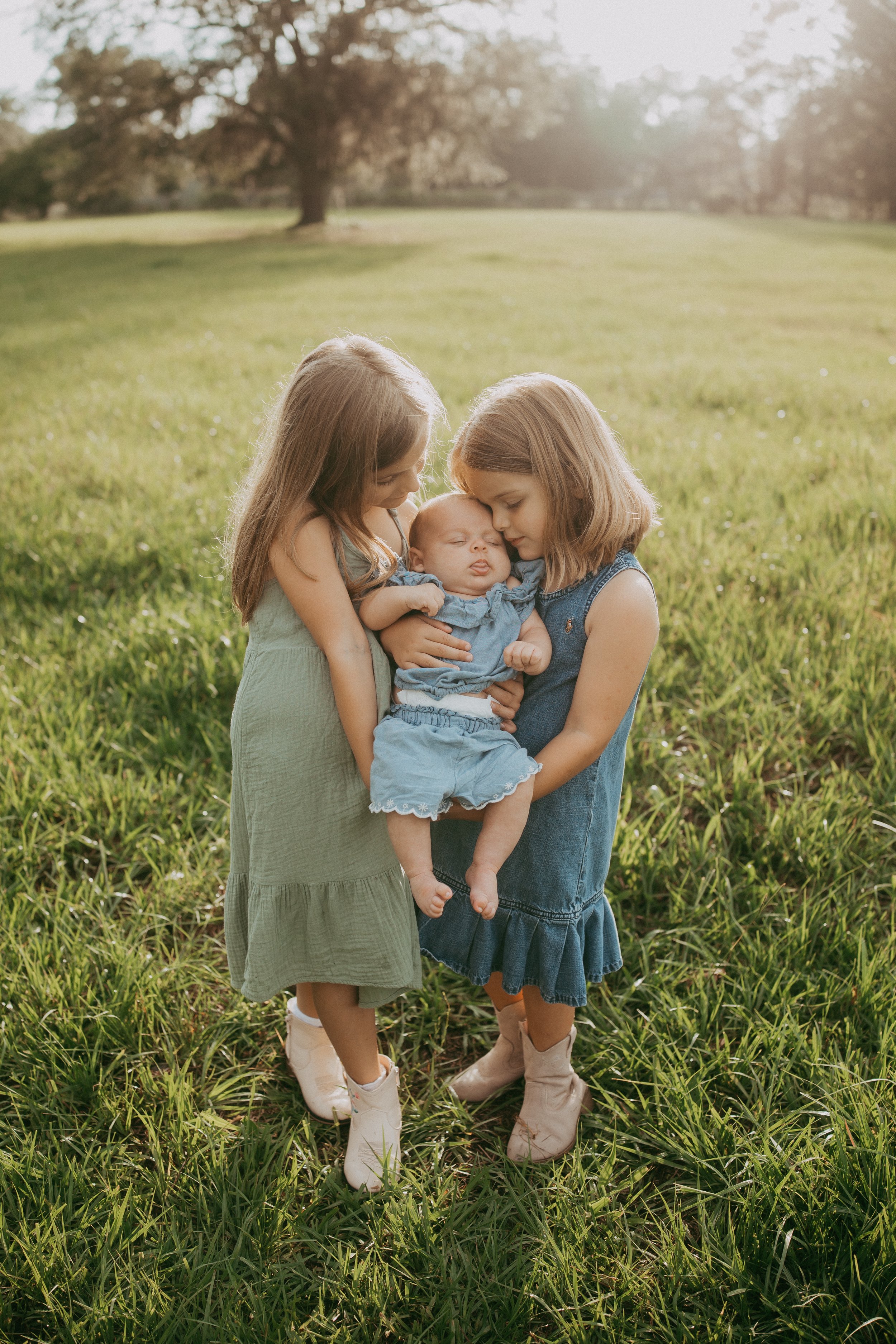 Two young sisters lovingly cradle their sleeping baby sibling in a sunlit grassy field, gently touching the newborn's cheeks in a heartwarming portrait celebrating the bond between sisters.
