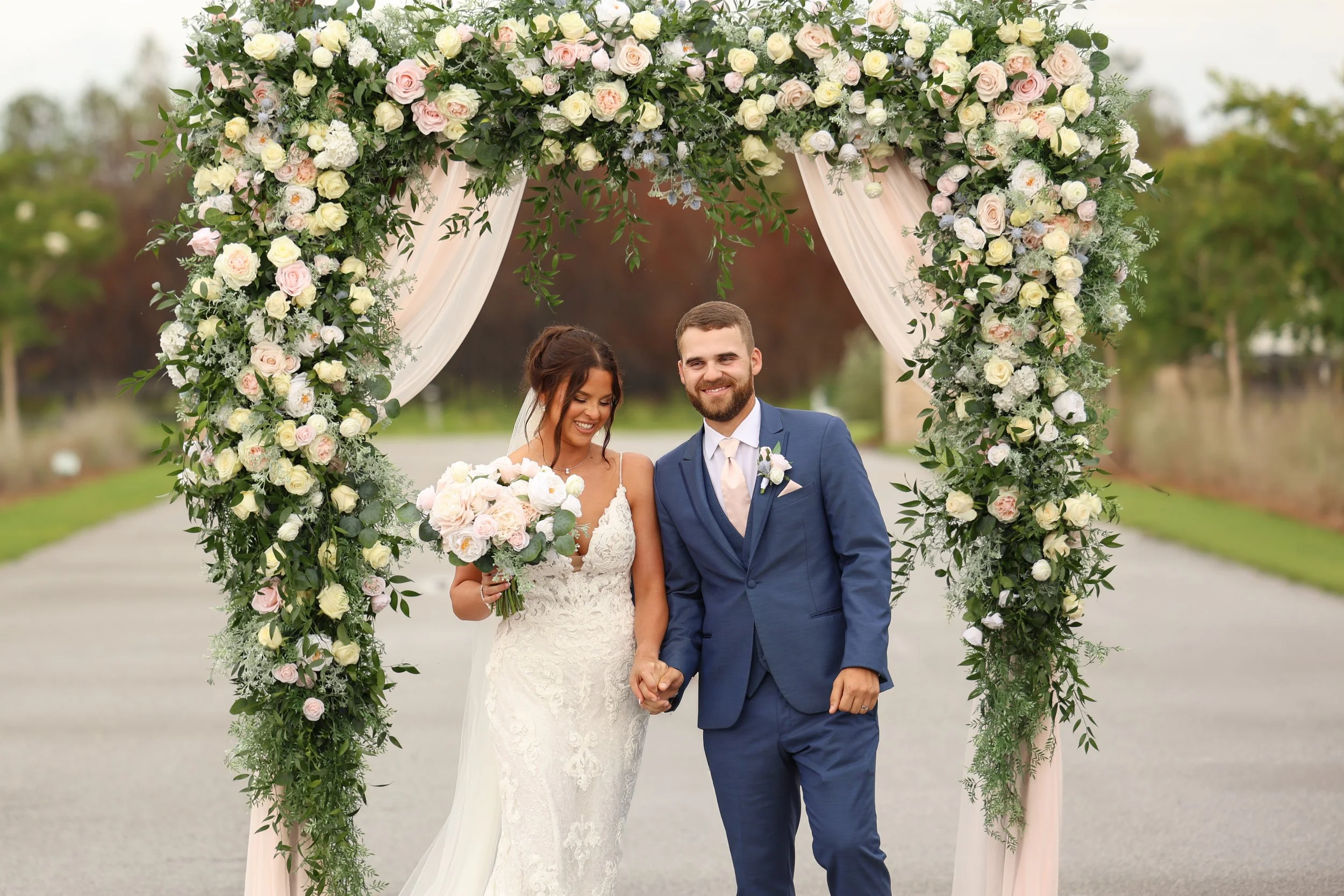 Newlyweds Emma and Dylan hold hands beneath a floral wedding arch covered in blush and ivory roses at Red River Estate in Waynesville, Georgia.