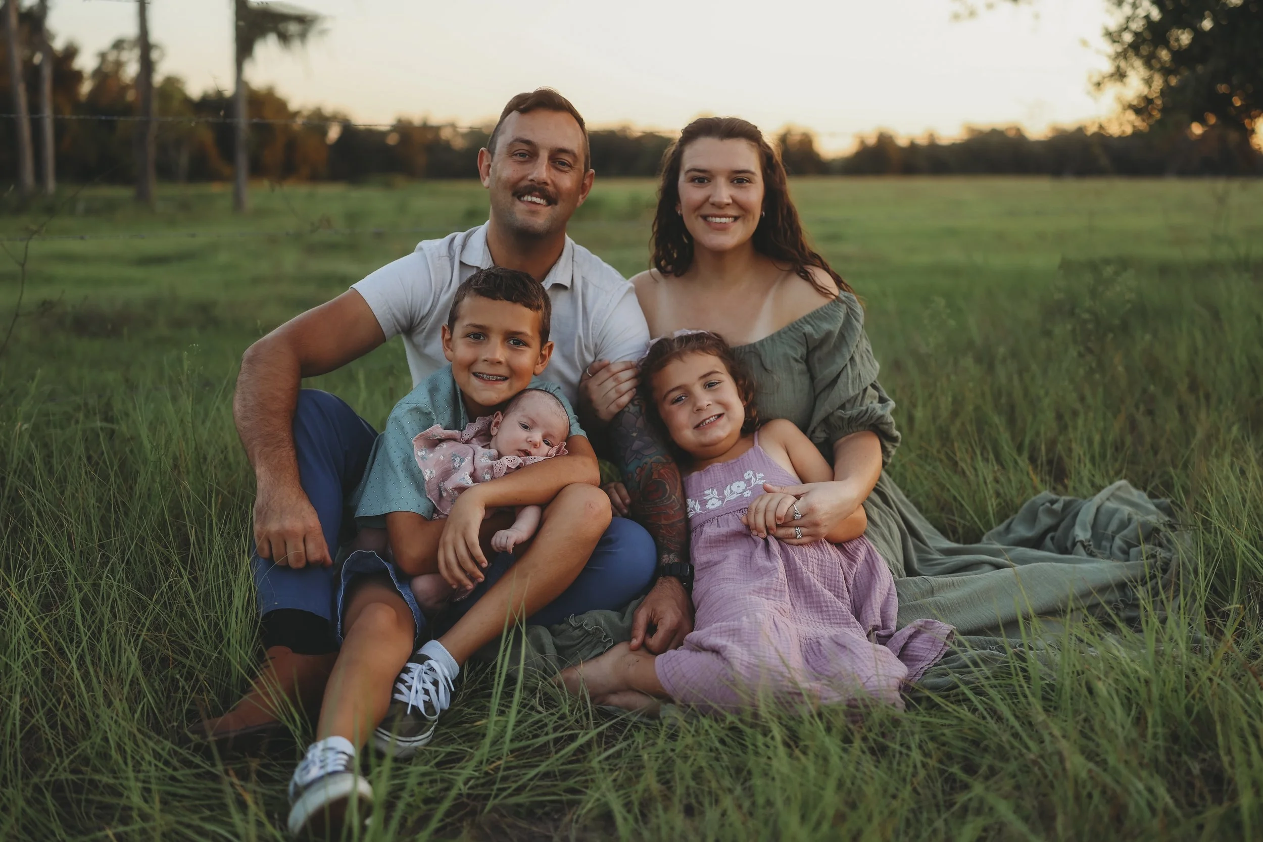 A family of five sits together in a grassy field at sunset, cuddled close as their oldest son gently holds his newborn baby sister while everyone smiles toward the camera.