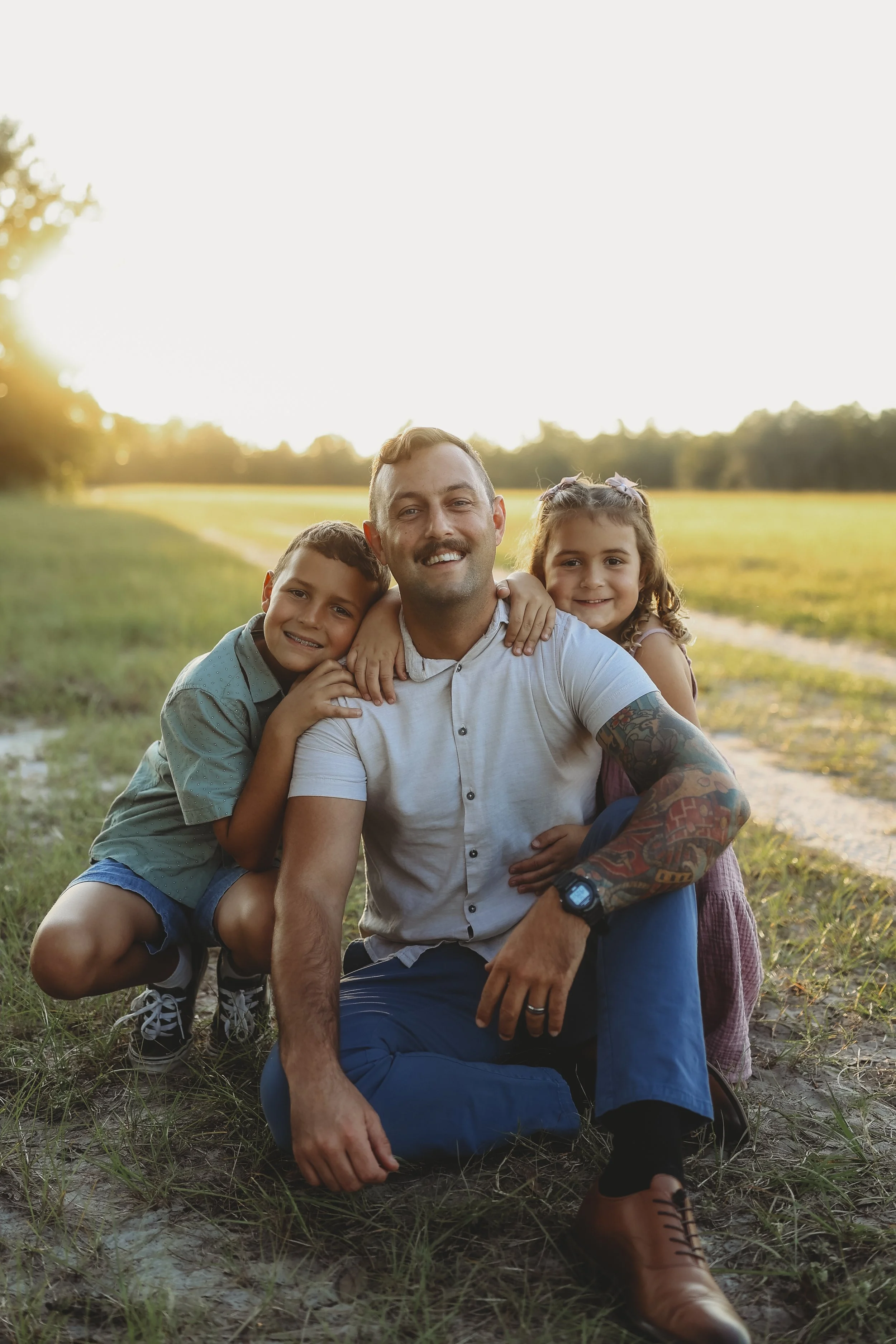 A father sits between his son and daughter as they wrap their arms around him, sharing a quiet moment together in a sunlit field at sunset.